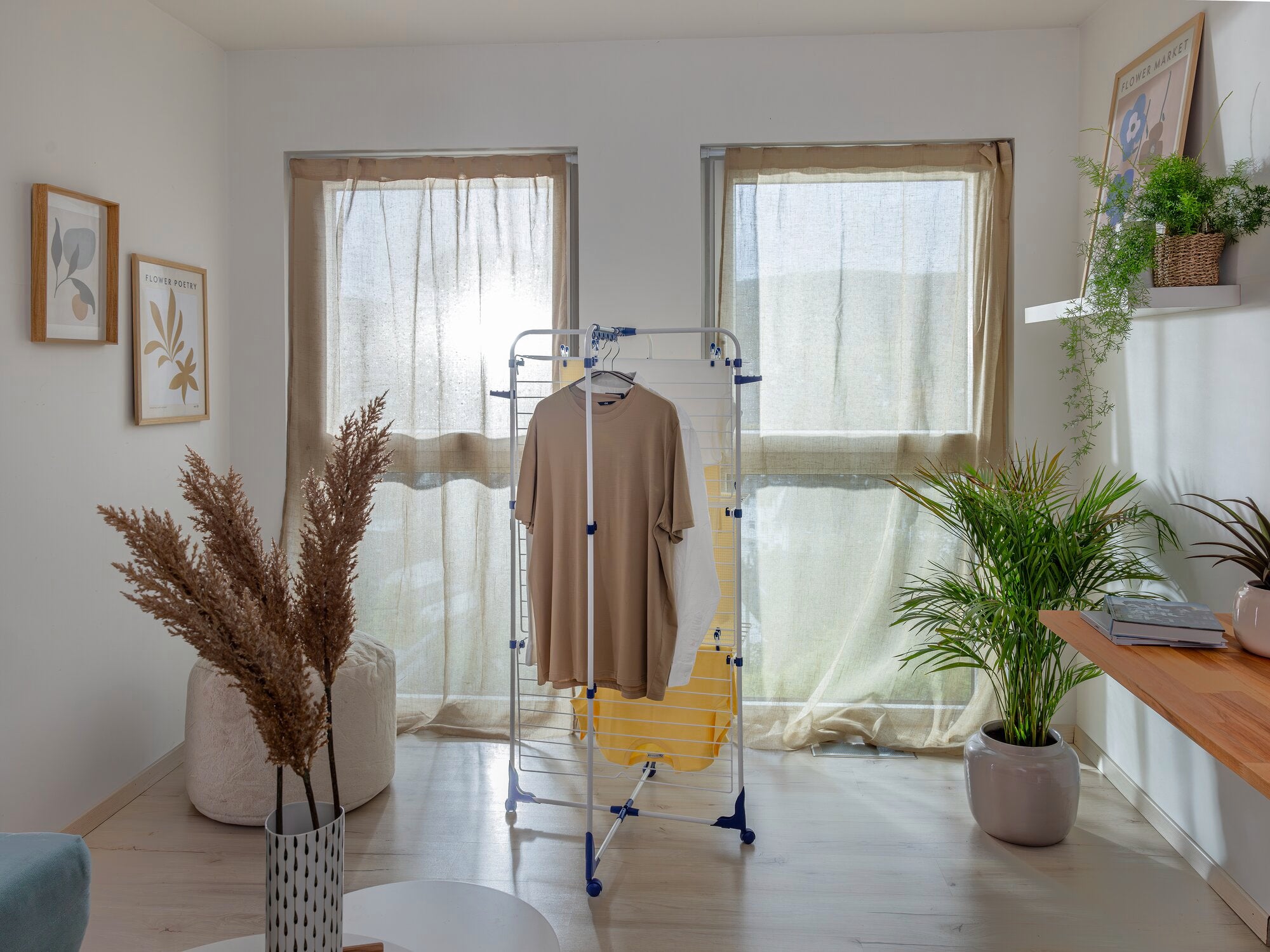 Een Classic Tower 340 toren droogtoren staat in een lichte kamer voor twee ramen. Er hangt een beige kledingstuk aan. Planten en decoraties omringen de wasdroger.