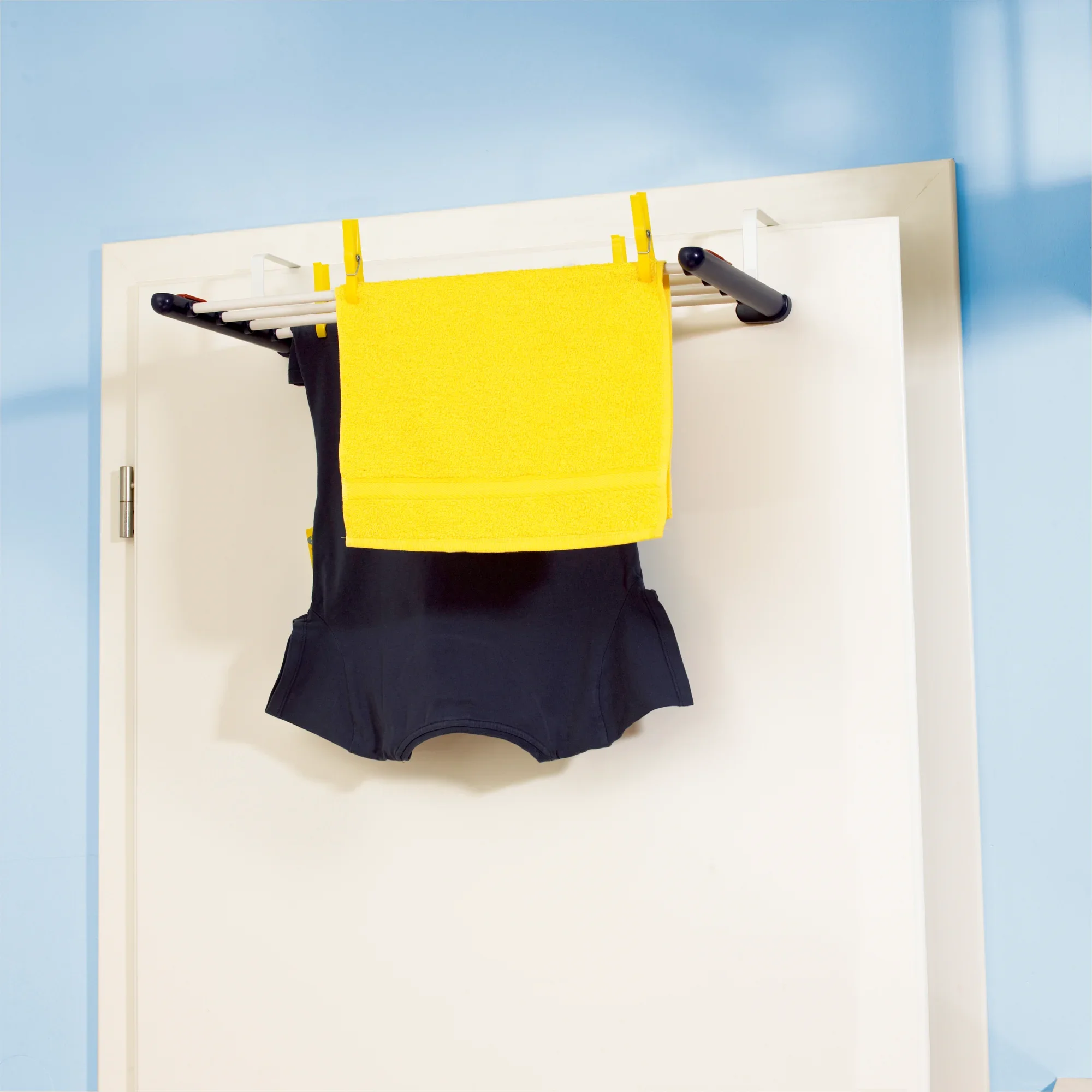 Een universele droger is gemonteerd op een deur. Er hangt een geel en een zwart kledingstuk in. De droger is compact en ruimtebesparend, ideaal voor kleine kamers.