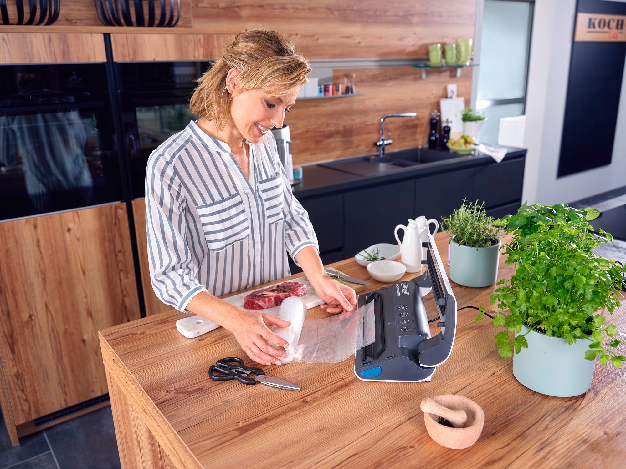 Eine Frau benutzt das Vakuumiergerät Vacu Power 300 in einer modernen Küche. Sie vakuumiert Fleisch, um es frisch zu halten. Auf der Arbeitsplatte stehen Kräuter und Küchenutensilien.