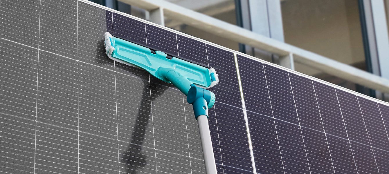 Ein tuerkisfarbener Wischer reinigt ein grosses Solarpanel, reflektiert das Sonnenlicht, vor einer modernen Glasfassade.