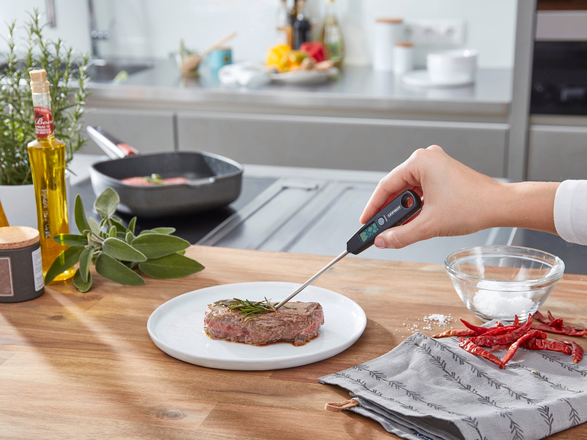 Een digitale universele thermometer wordt gebruikt om de temperatuur van een steak op een bord te meten. Op de achtergrond is een moderne keuken met verse kruiden en specerijen te zien.