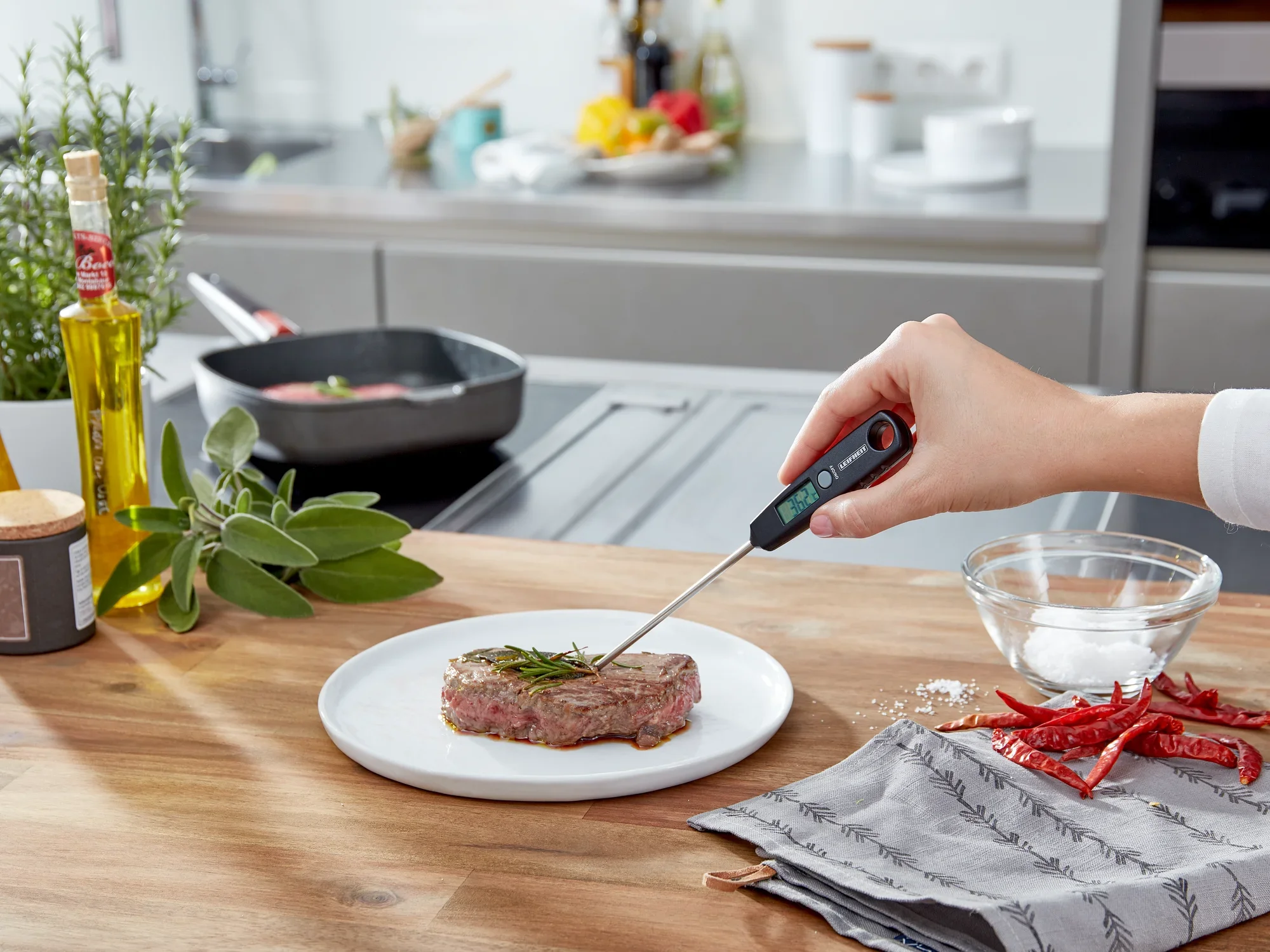 Cyfrowy termometr uniwersalny służy do pomiaru temperatury steku na talerzu. W tle widać nowoczesną kuchnię ze świeżymi ziołami i przyprawami.