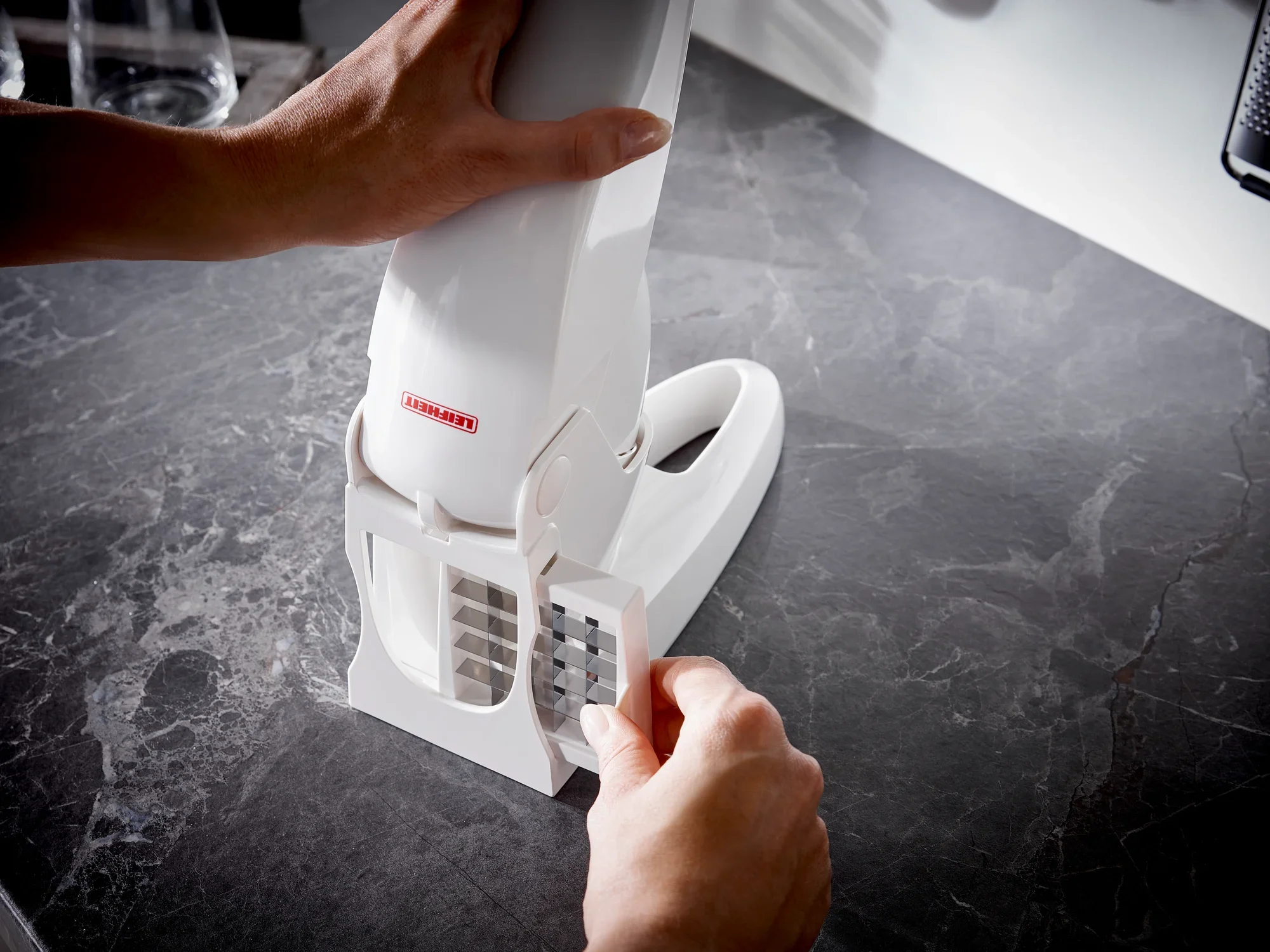 Een witte frietsnijder wordt gebruikt op een donkergekleurd keukenblad. Eén persoon houdt de frietsnijder stevig vast terwijl hij het mes erin steekt. Ideaal voor gelijkmatige frietjes.