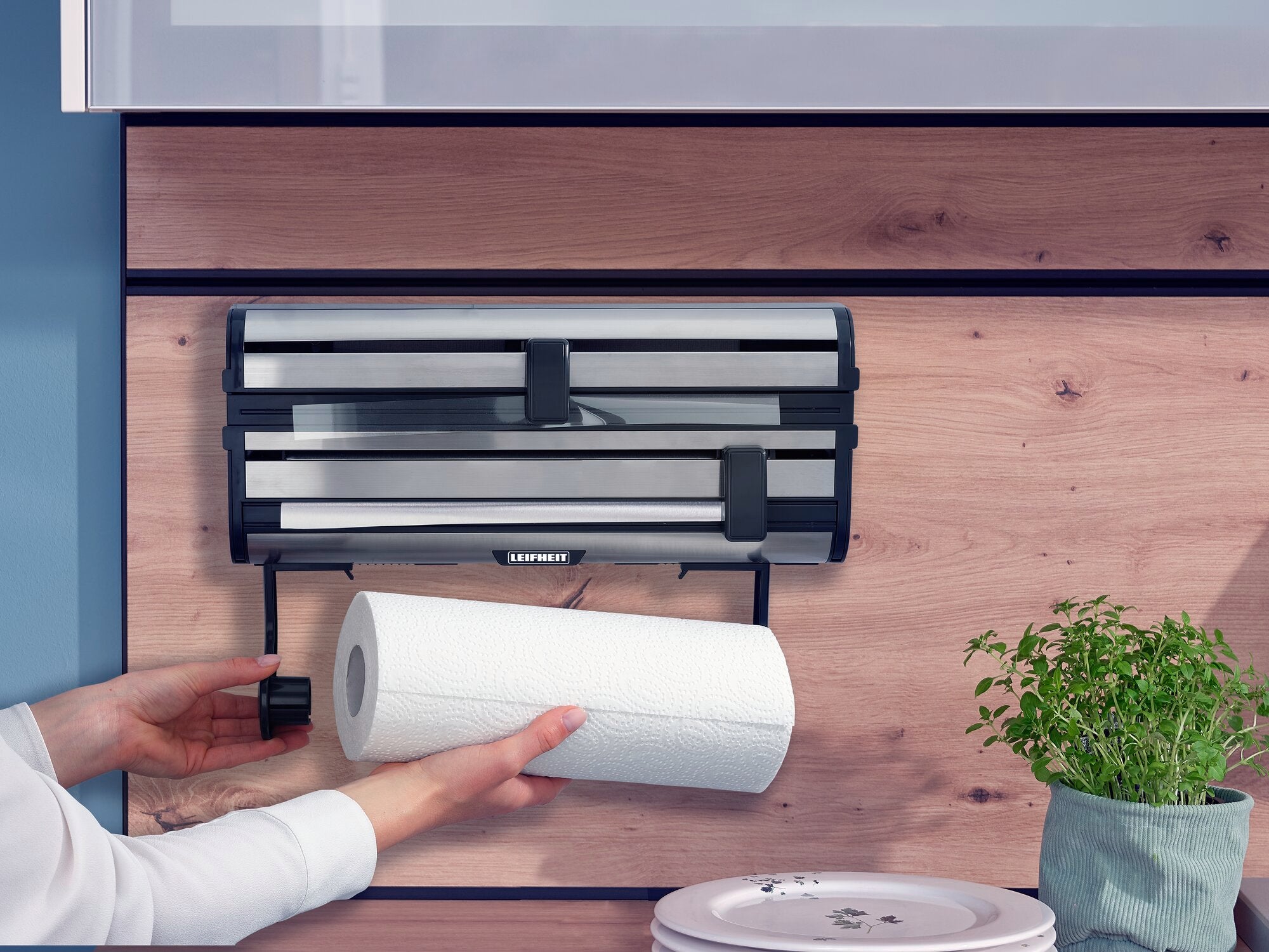 Parat Royal wall-mounted roll holder mounted on a wooden wall. A hand reaches for a kitchen roll hanging in the holder. Next to it is a pot of fresh herbs.