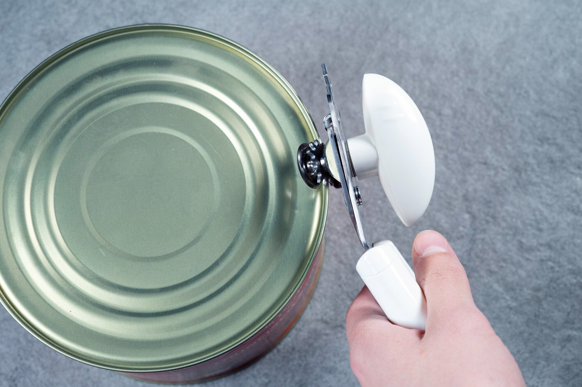 Ouvre-boîte Exact en action : une main utilise l'ouvre-boîte blanc pour ouvrir le couvercle d'une grande boîte de conserve sur un fond gris.