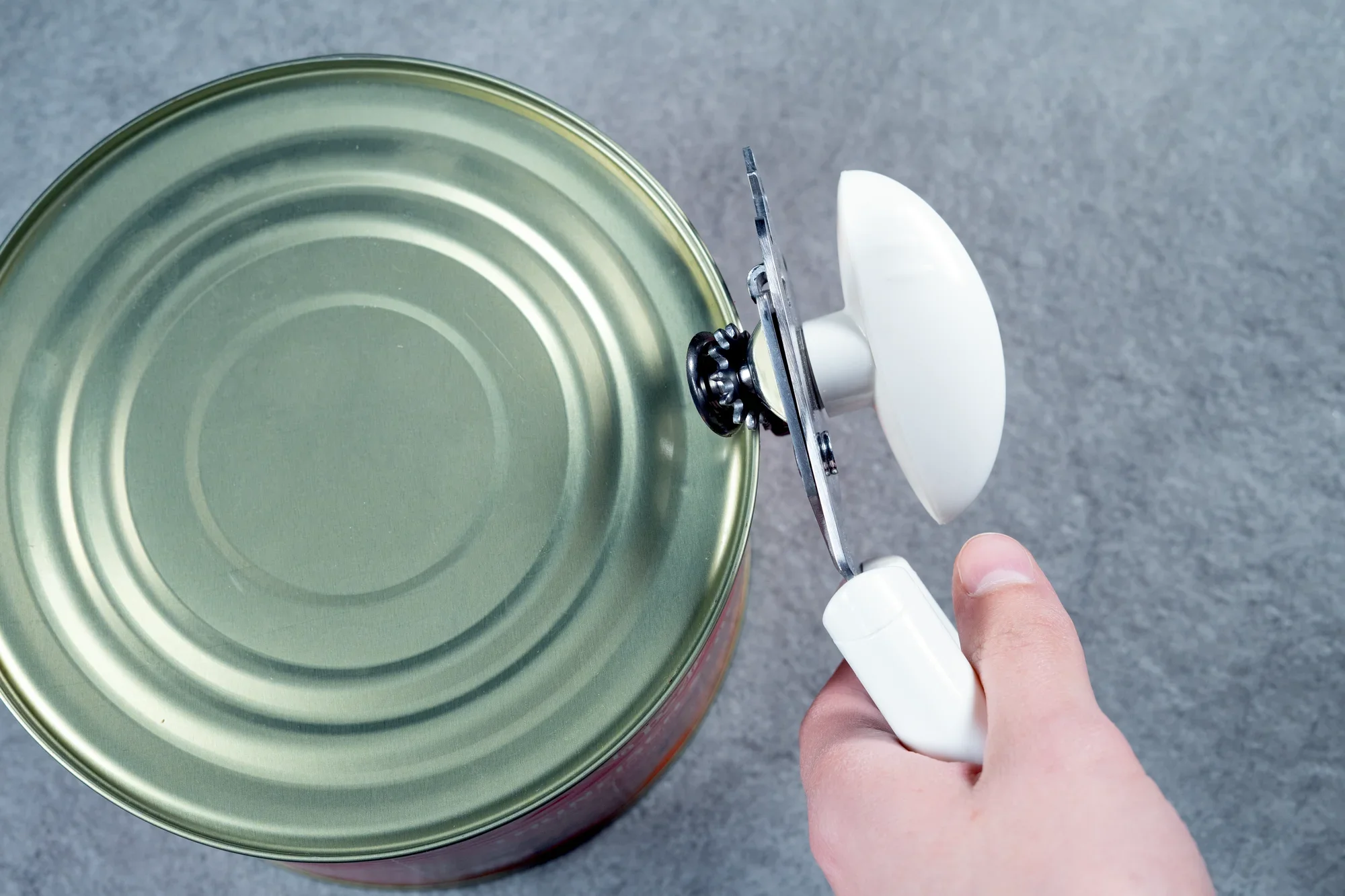 Otwieracz do puszek Exact w użyciu: Jedna ręka używa białego otwieracza do puszek, aby otworzyć pokrywkę dużej puszki na szarym tle.