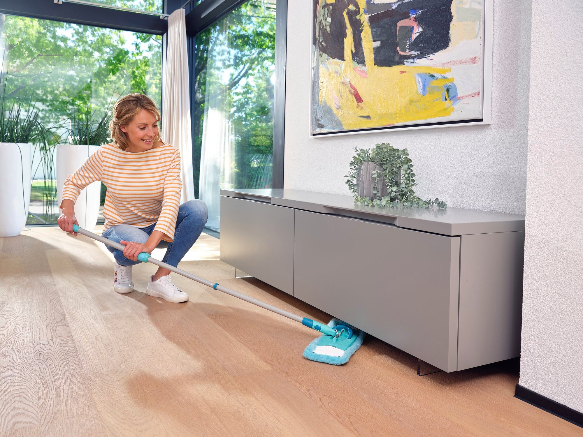 A woman cleans the wooden floor in a modern living room with the CLEAN TWIST mop. The mop easily reaches hard-to-reach places under a low cabinet.