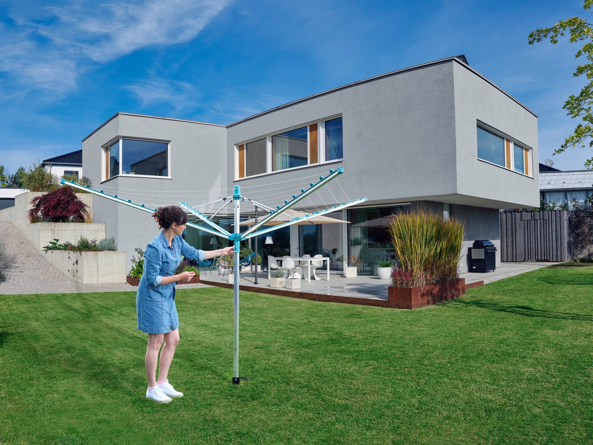 Eine Frau benutzt den Wäscheschirm Linomatic 500 Easy im Garten eines modernen Hauses. Der Schirm ist geöffnet und bietet viel Platz zum Trocknen von Kleidung.