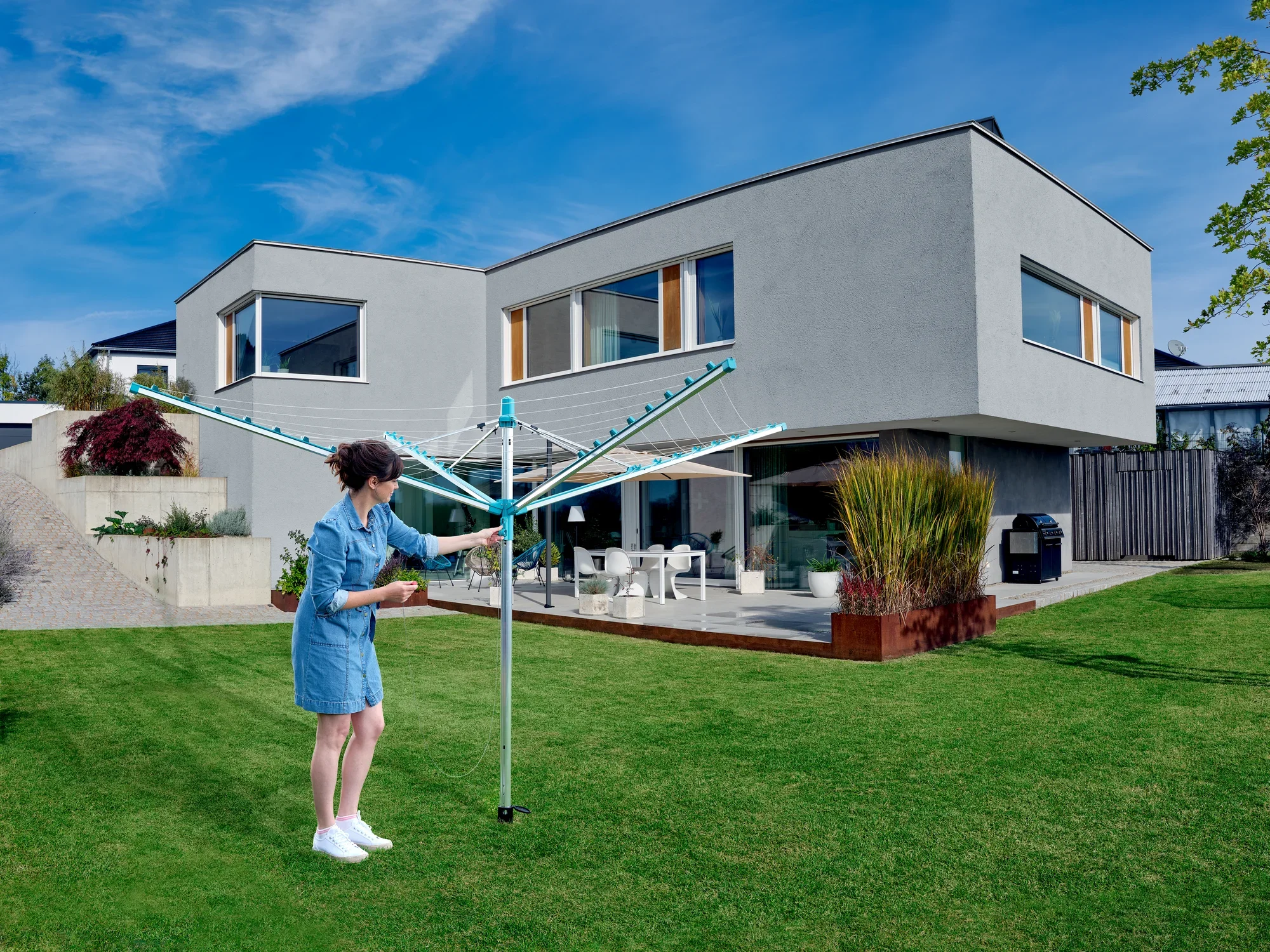 Kobieta korzysta z parasola do prania Linomatic 500 Easy w ogrodzie nowoczesnego domu. Parasol jest otwarty i oferuje dużo miejsca do suszenia ubrań.