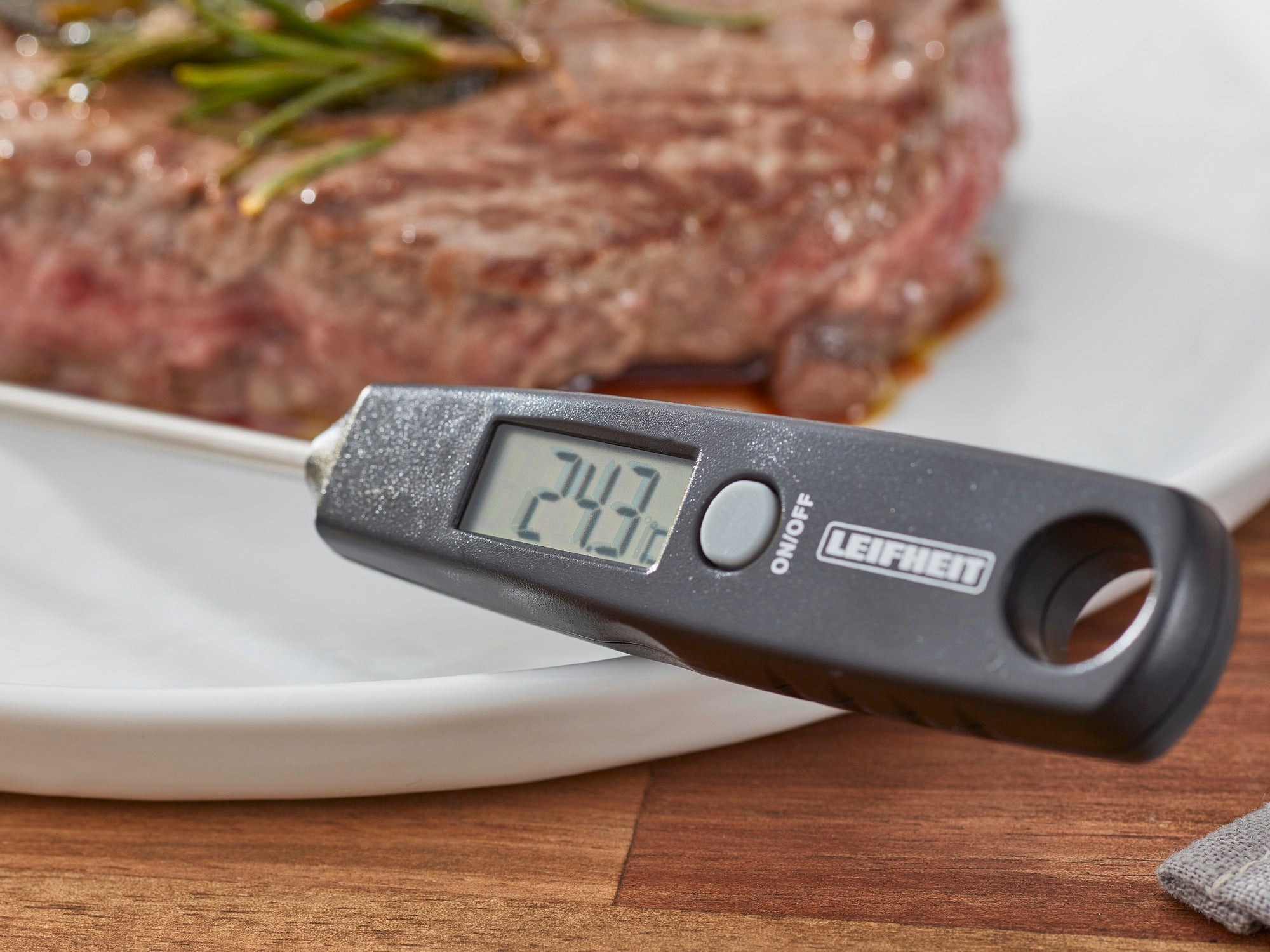 Ein digitales Kuechenthermometer zeigt 24,3 Grad Celsius an, waehrend es neben einem saftigen, mit Rosmarin garnierten Steak liegt.