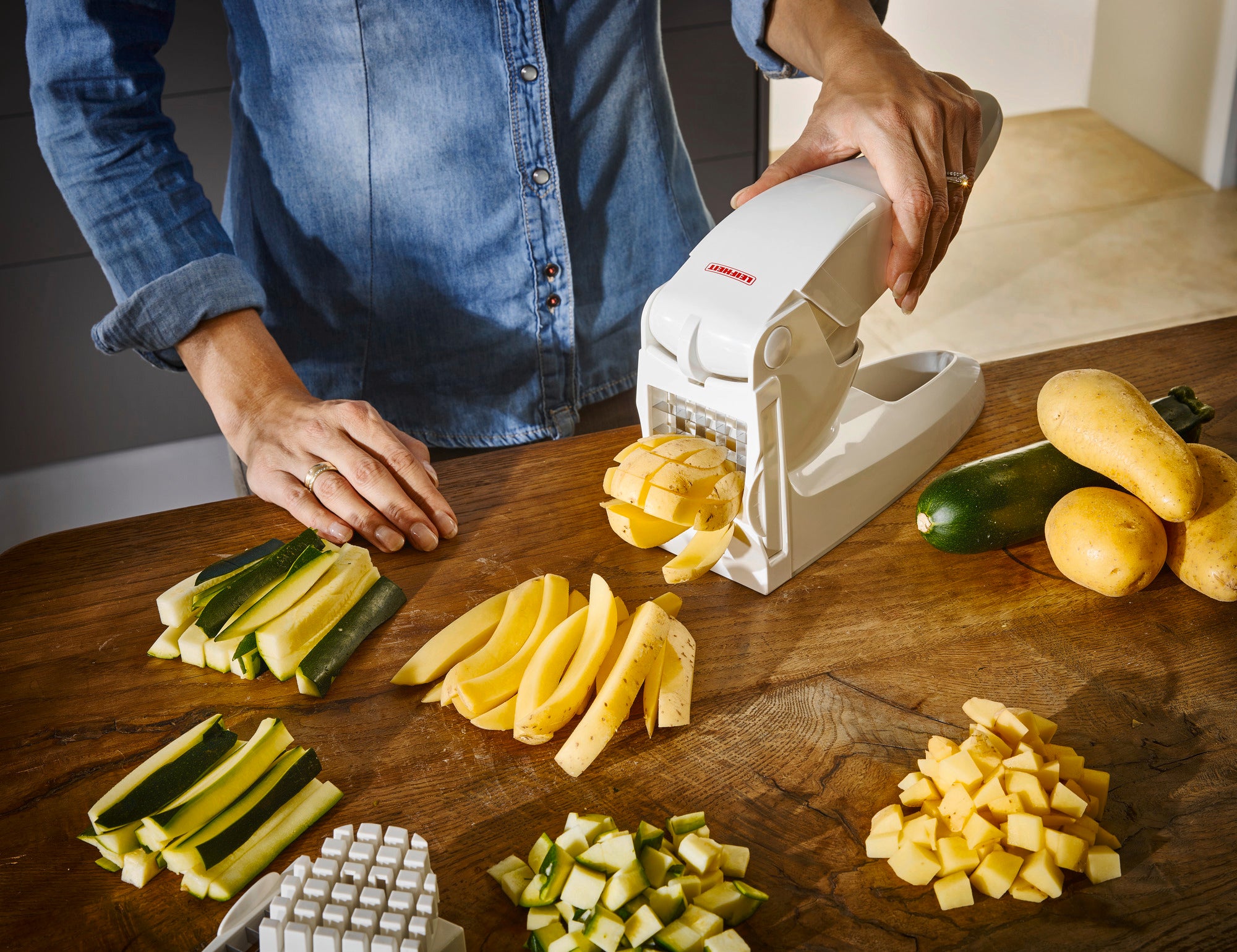 Eine Person benutzt einen Pommes-Frites-Schneider, um Gemüse in gleichmäßige Stifte zu schneiden. Auf dem Tisch liegen geschnittene Zucchini, Kartoffeln und andere Gemüsesorten.