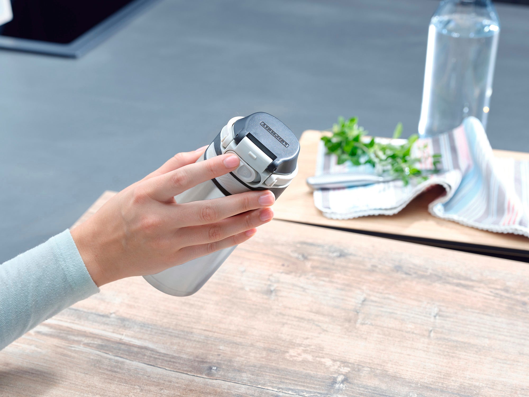 Flip 350ml insulated mug in silver is held by one hand. The mug has a practical lid. A tea towel and herbs can be seen in the background.