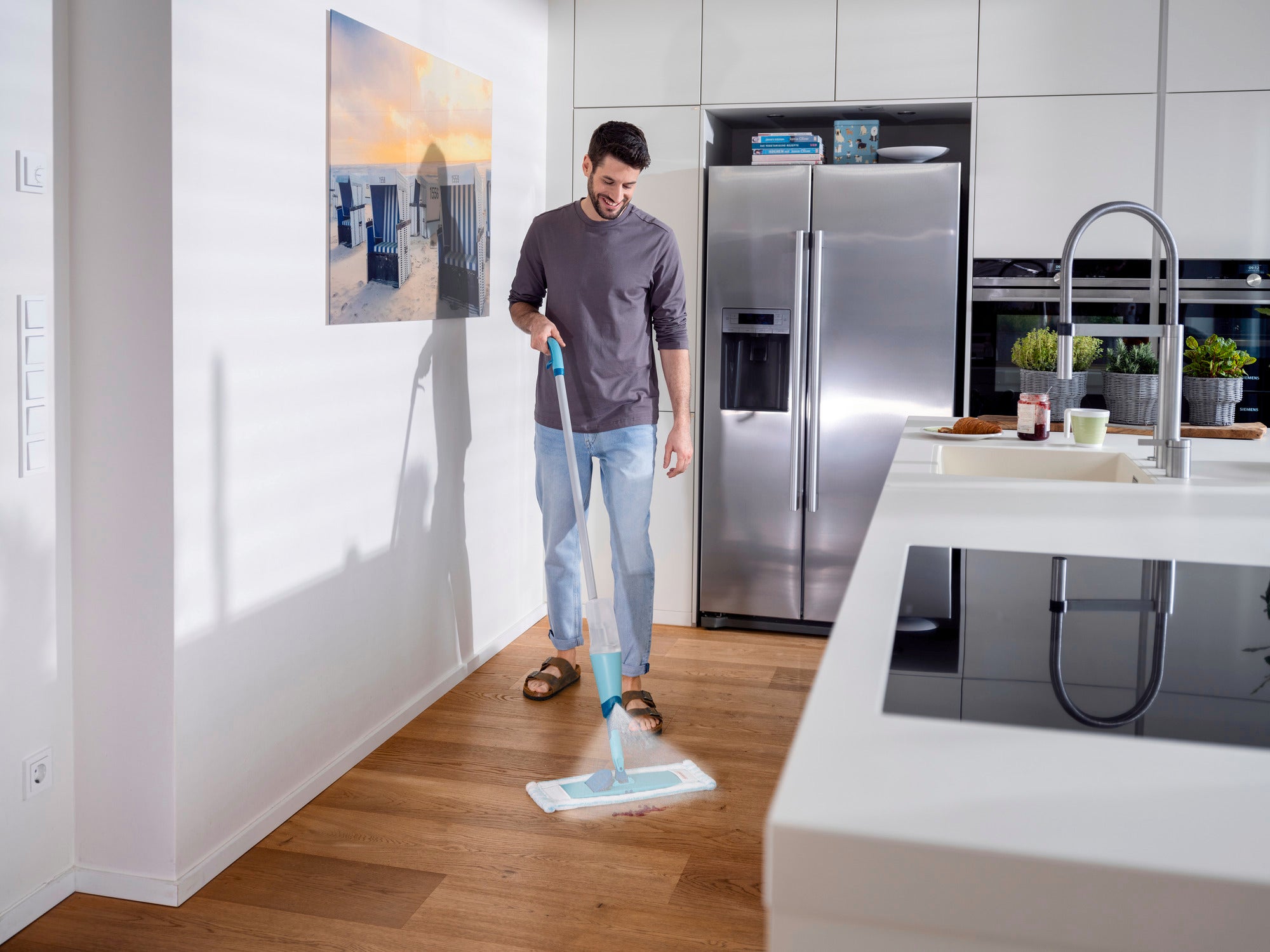 Een man maakt de houten vloer van een moderne keuken schoon met de Easy Spray XL spraymop. De keuken is licht en stijlvol, met roestvrijstalen apparatuur en een grote koelkast.
