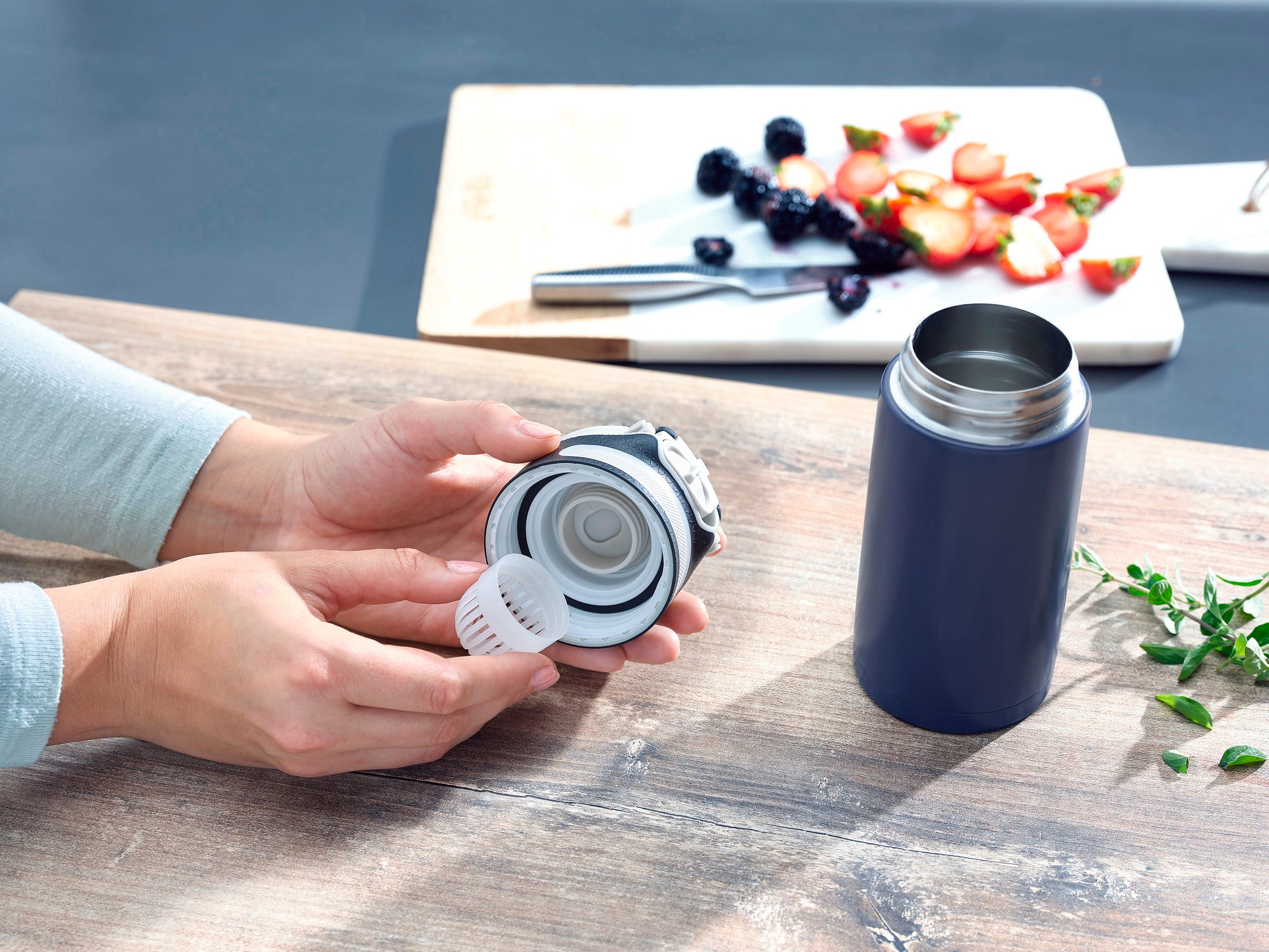 Flip 350 ml insulated mug in dark blue on a table. A person is holding the lid with integrated strainer. In the background is a cutting board with fruit. Ideal for on the go.
