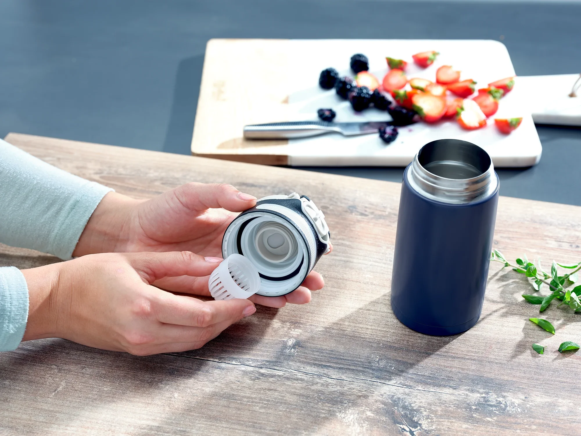 Kubek próżniowy Flip 350 ml w kolorze ciemnoniebieskim na stole. Osoba trzyma pokrywkę z wbudowanym sitkiem. W tle deska do krojenia z owocami. Idealny do podróży.