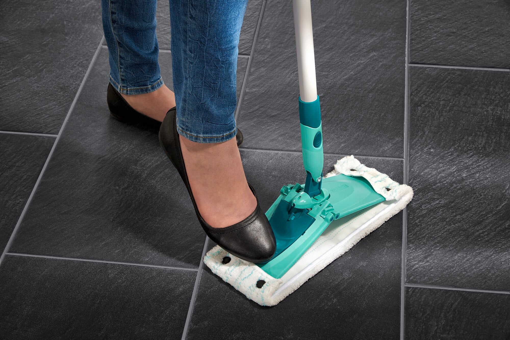Une personne appuie avec son pied sur le balai Combi M pour détacher la housse. Le balai repose sur un carrelage foncé et démontre sa praticité au quotidien.