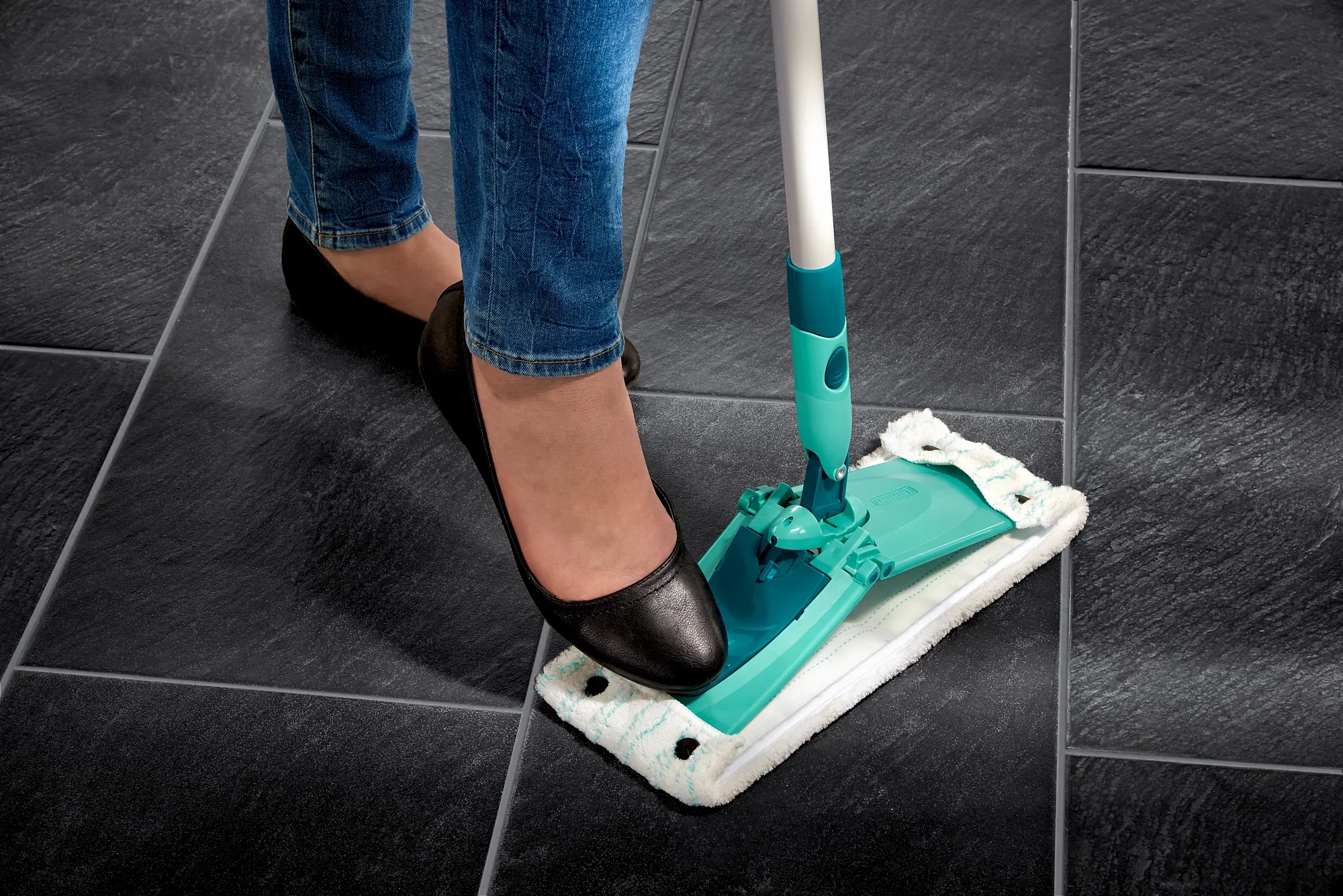 A person presses down on the Combi M floor mop with their foot to release the mop head. The mop lies on dark tiles, demonstrating its practicality in everyday use.