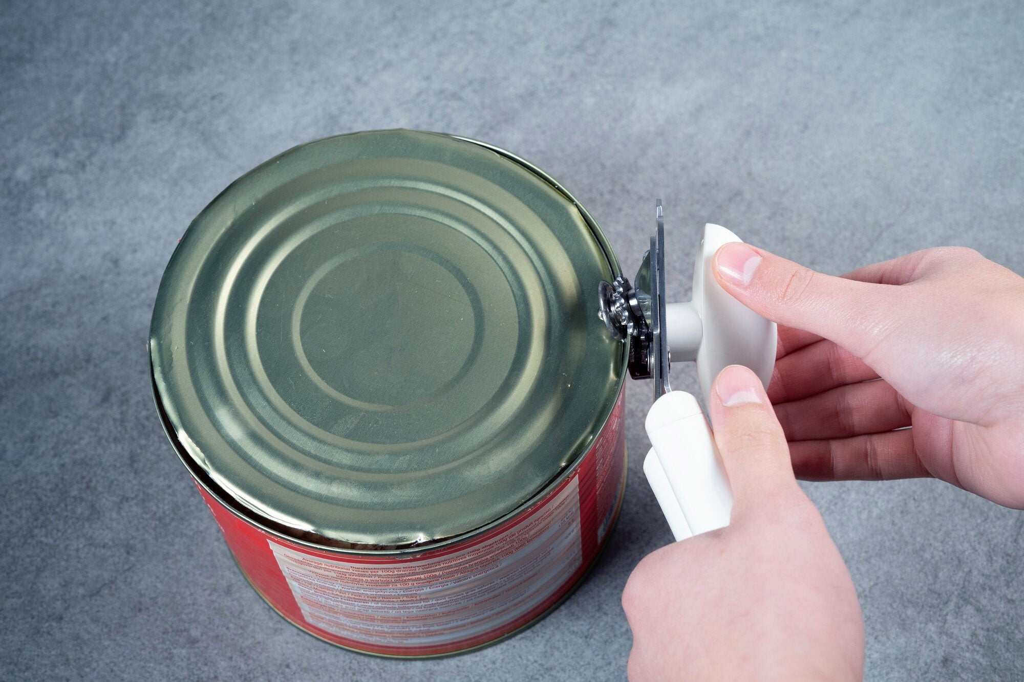 Une personne ouvre une grande boite de conserve en metal avec un ouvre-boite manuel sur une surface grise.