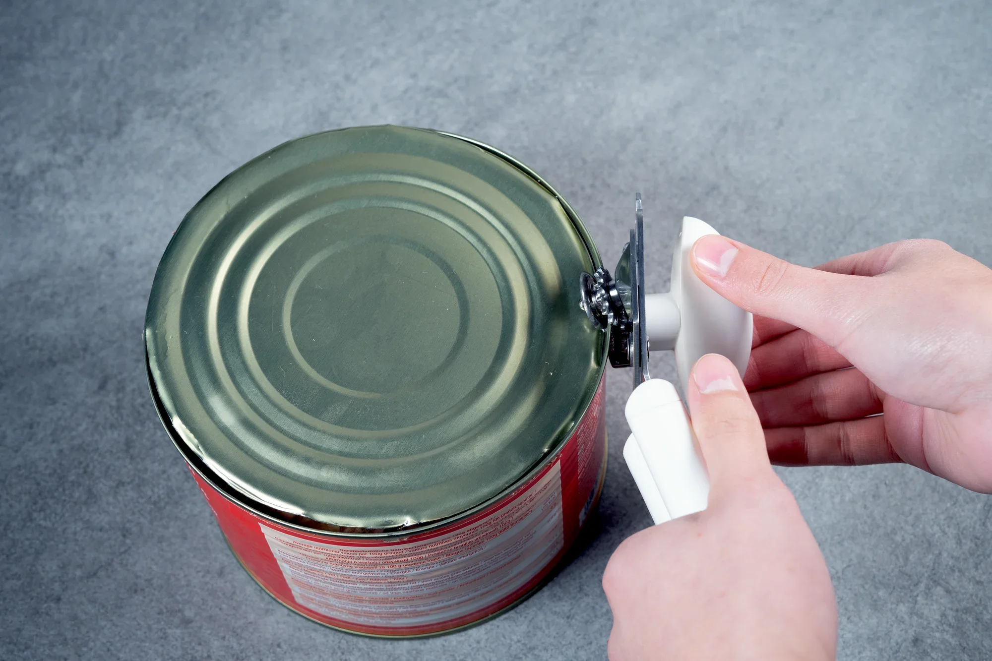 Osoba używa otwieracza do puszek Exact, aby otworzyć dużą puszkę na szarej powierzchni. Otwieracz ma białą rączkę i zapewnia precyzyjne cięcie.