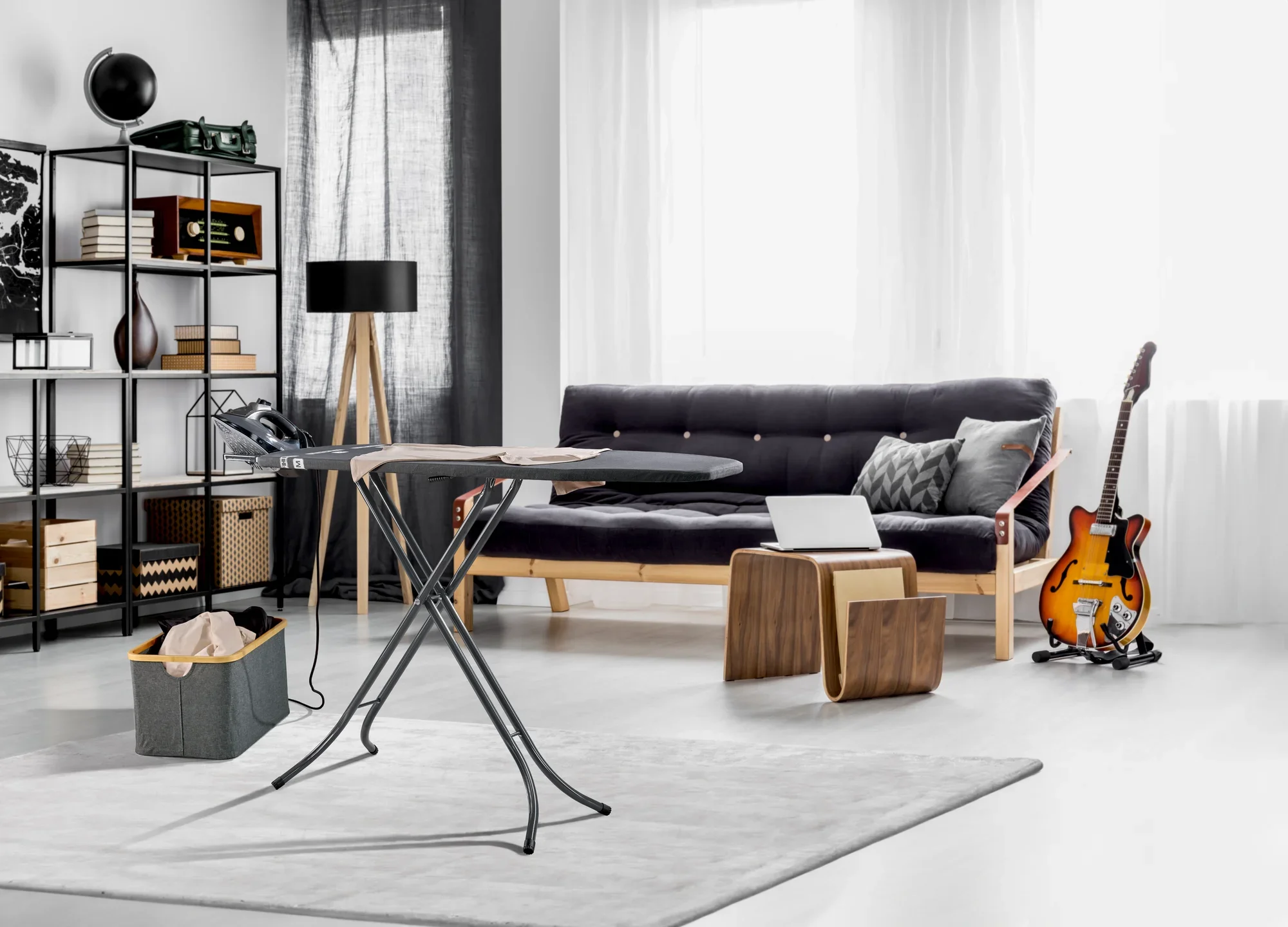 Classic M strijkplank in een moderne woonkamer met een grijze bank, houten meubels en een elektrische gitaar. De tafel staat op een lichtgekleurd tapijt en is klaar voor gebruik.