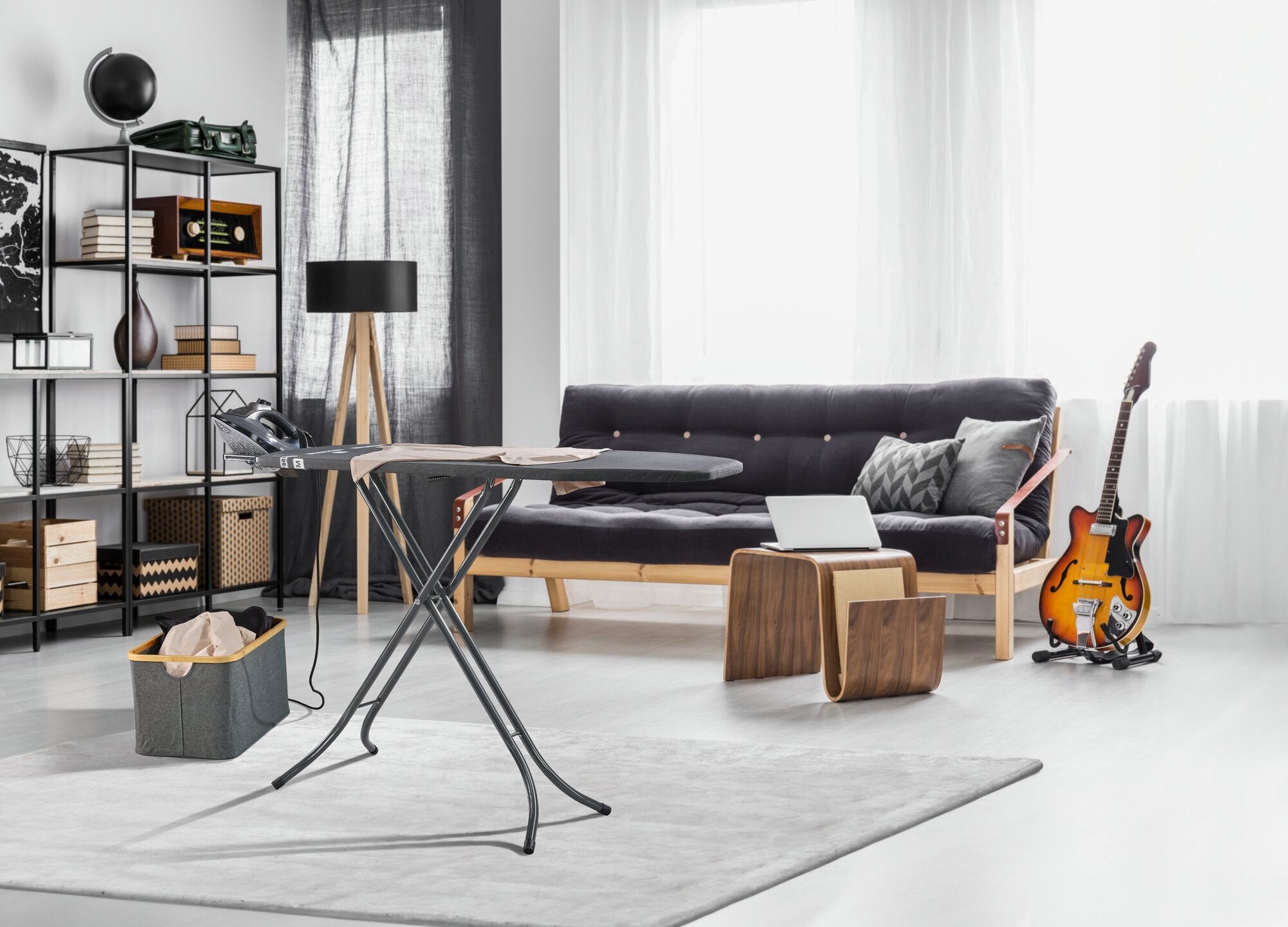 Classic M strijkplank in een moderne woonkamer met een grijze bank, houten meubels en een elektrische gitaar. De tafel staat op een lichtgekleurd tapijt en is klaar voor gebruik.