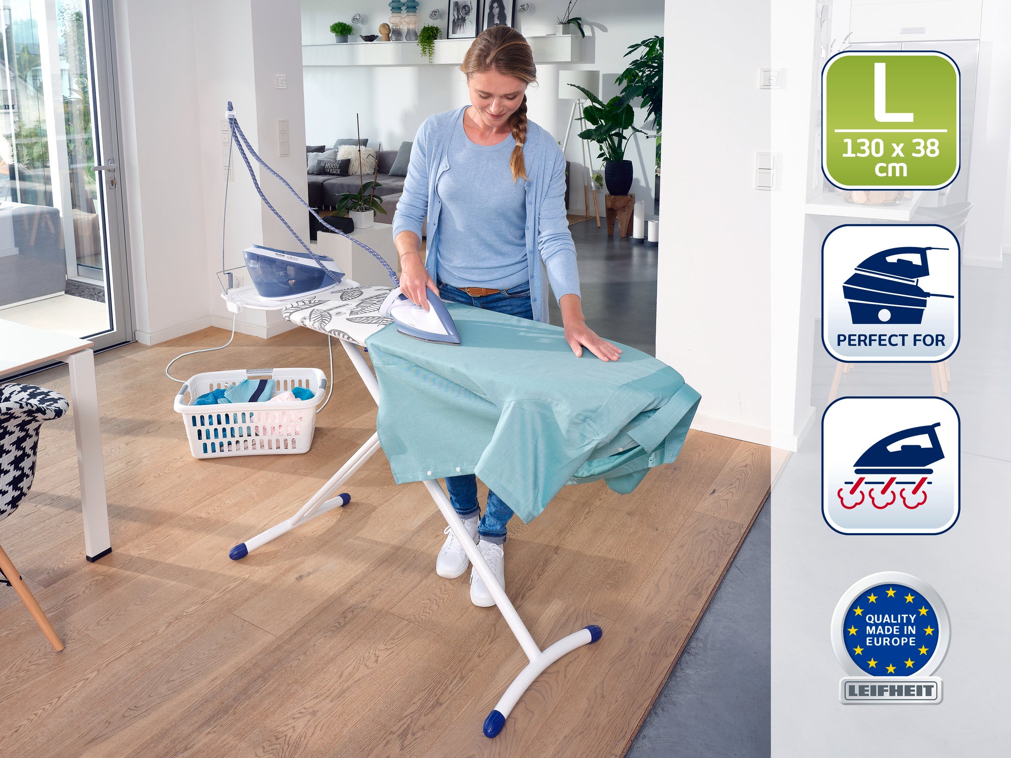 A woman irons a green cloth on an ironing table in a modern, bright room. The table measures 130 x 38 cm and is equipped with various features that make ironing easier.