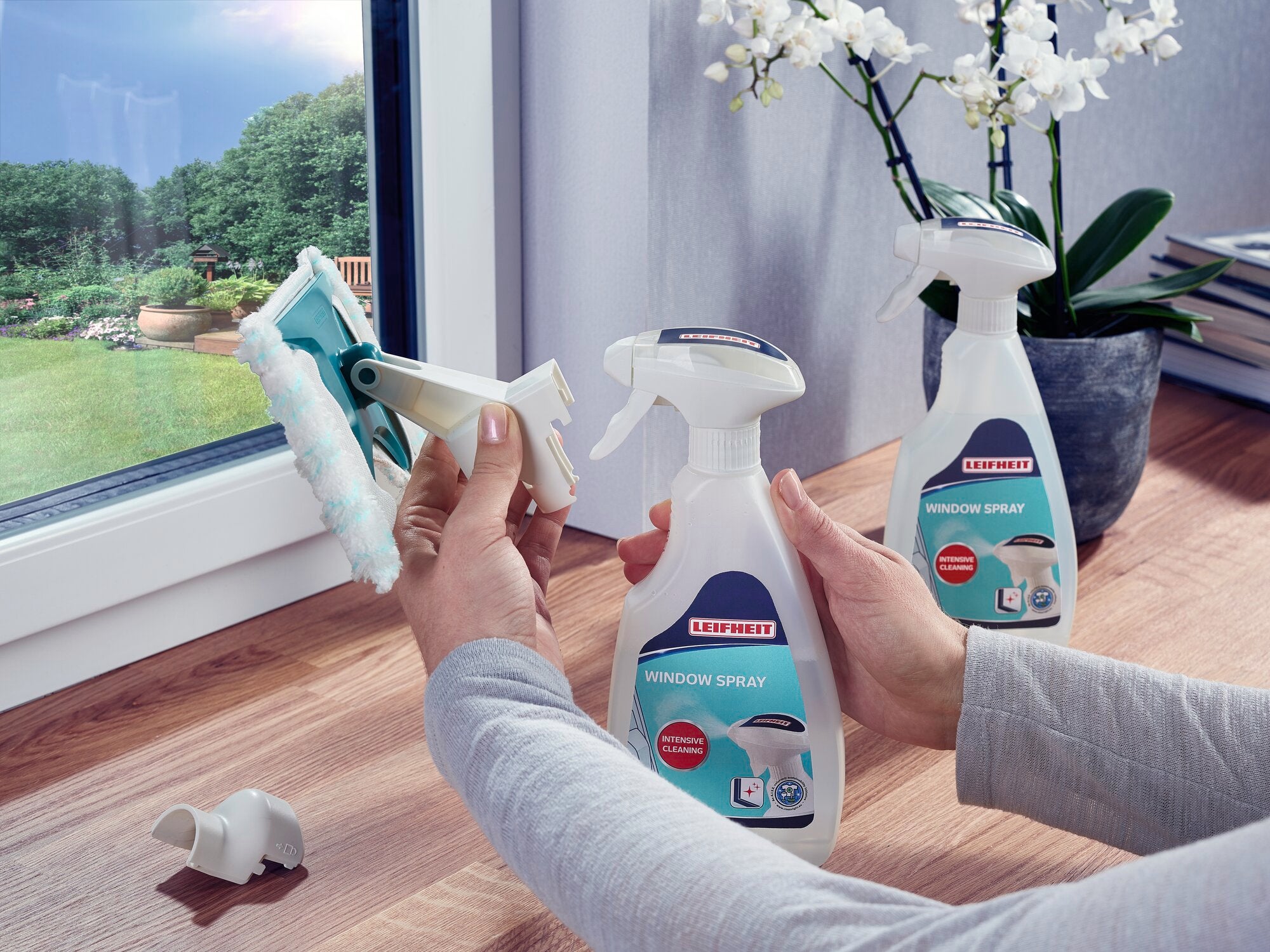 Zwei Hände verwenden den Leifheit Window Spray Cleaner, um ein Fenster zu reinigen. Im Hintergrund sind eine Orchidee und Bücher auf einem Holztisch zu sehen.