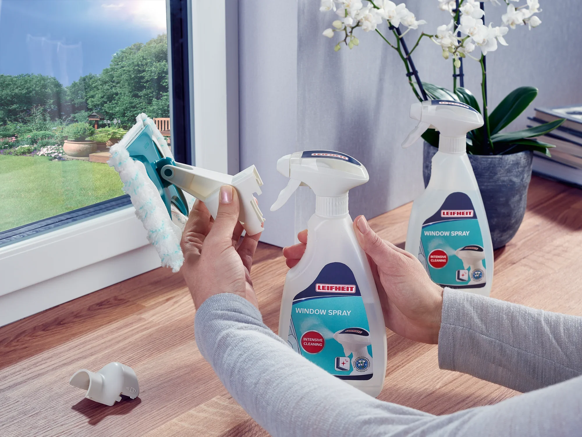 Dwie ręce używają środka do czyszczenia okien Leifheit Window Spray Cleaner do czyszczenia okna. W tle widać orchideę i książki na drewnianym stole.