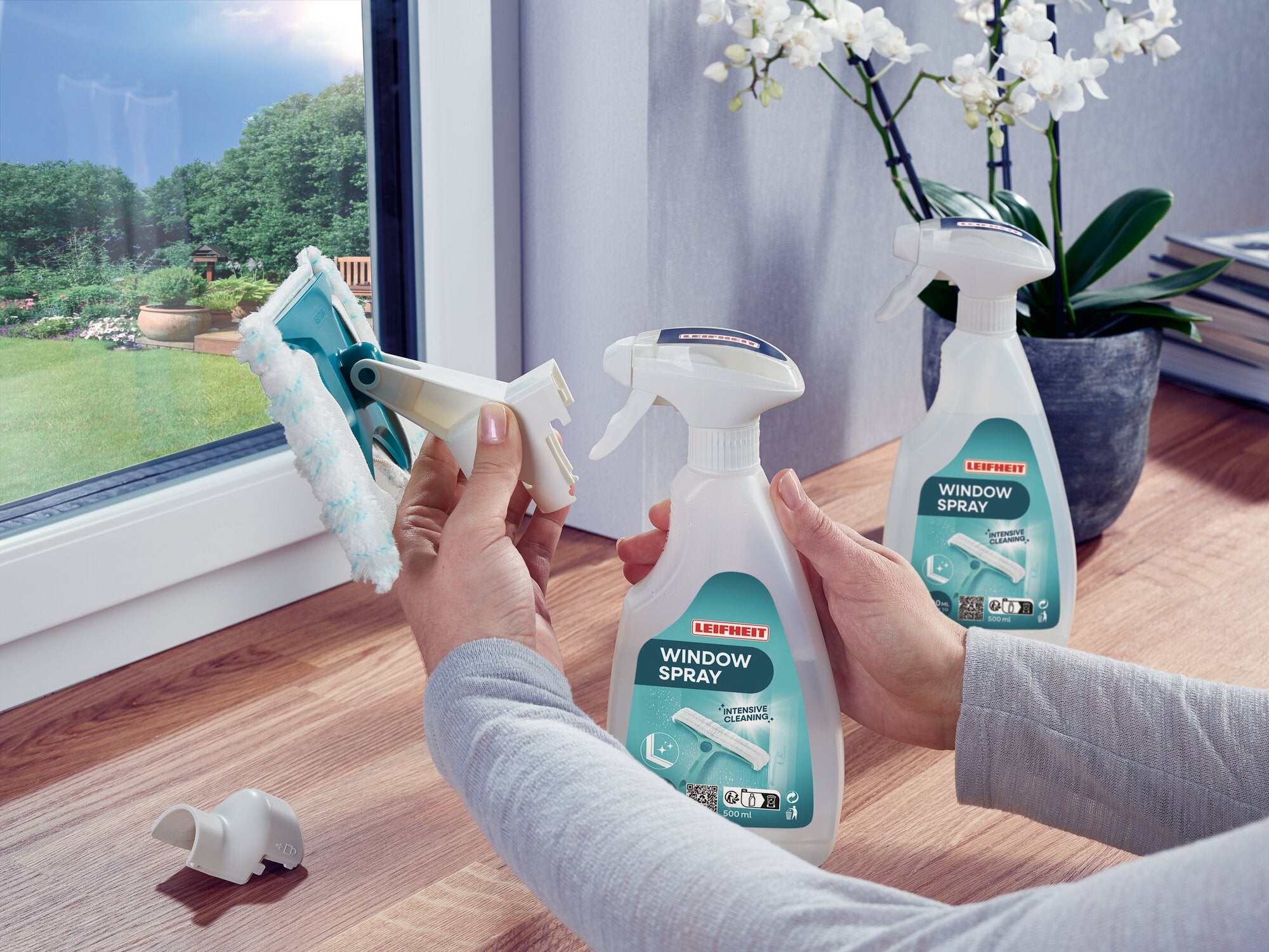 Zwei Hände verwenden den Leifheit Window Spray Cleaner, um ein Fenster zu reinigen. Im Hintergrund sind eine Orchidee und Bücher auf einem Holztisch zu sehen.