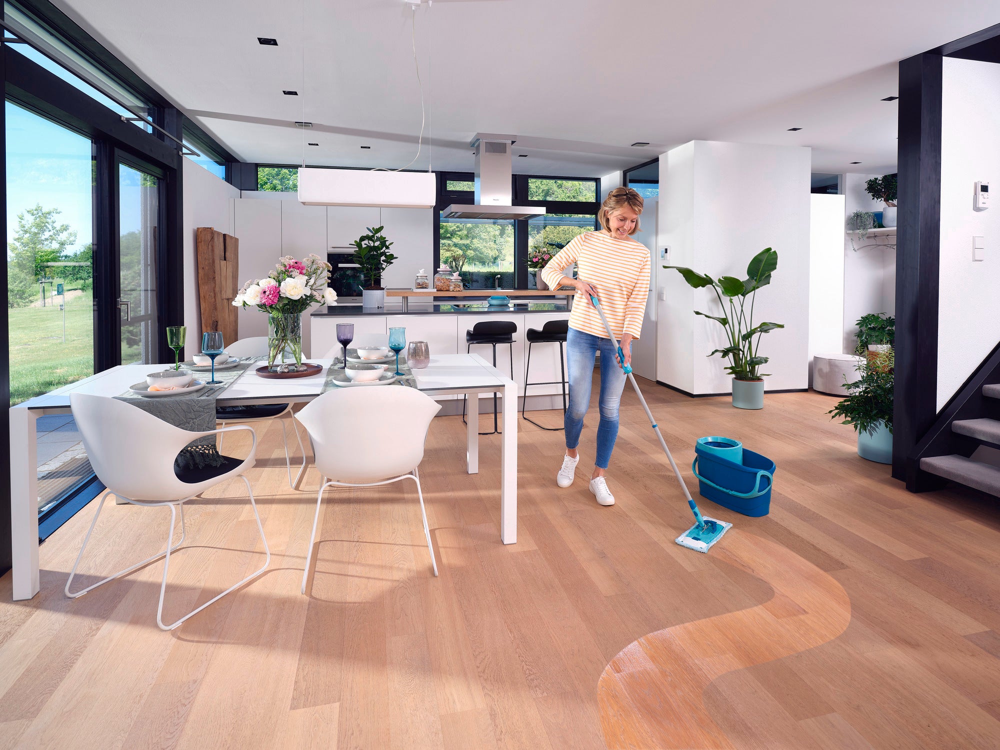 Een vrouw reinigt de houten vloer in een moderne, lichte woonkamer met de CLEAN TWIST M Ergo Set. De kamer is stijlvol ingericht met een eettafel, stoelen en planten.