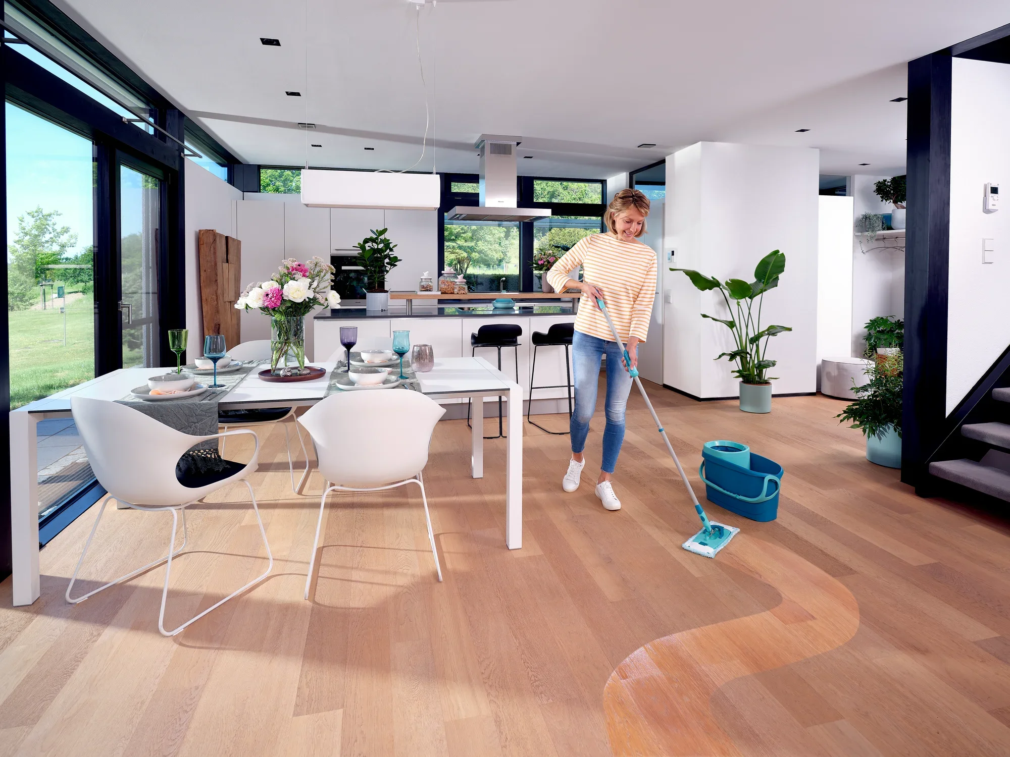 Une femme nettoie le sol en bois d'un salon moderne et lumineux avec le kit CLEAN TWIST M Ergo. La pièce est aménagée avec style, avec une table à manger, des chaises et des plantes.