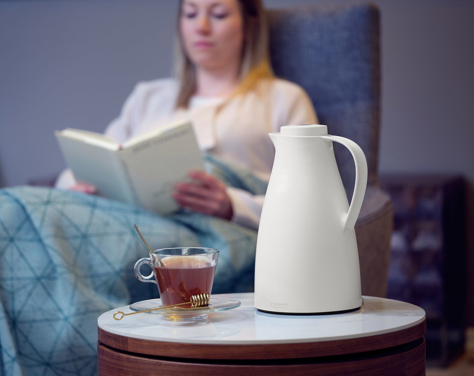Eine weiße Isolierkanne namens Harmonic steht auf einem runden Tisch neben einer Tasse Tee. Im Hintergrund sitzt eine Frau entspannt auf einem Sessel und liest ein Buch.