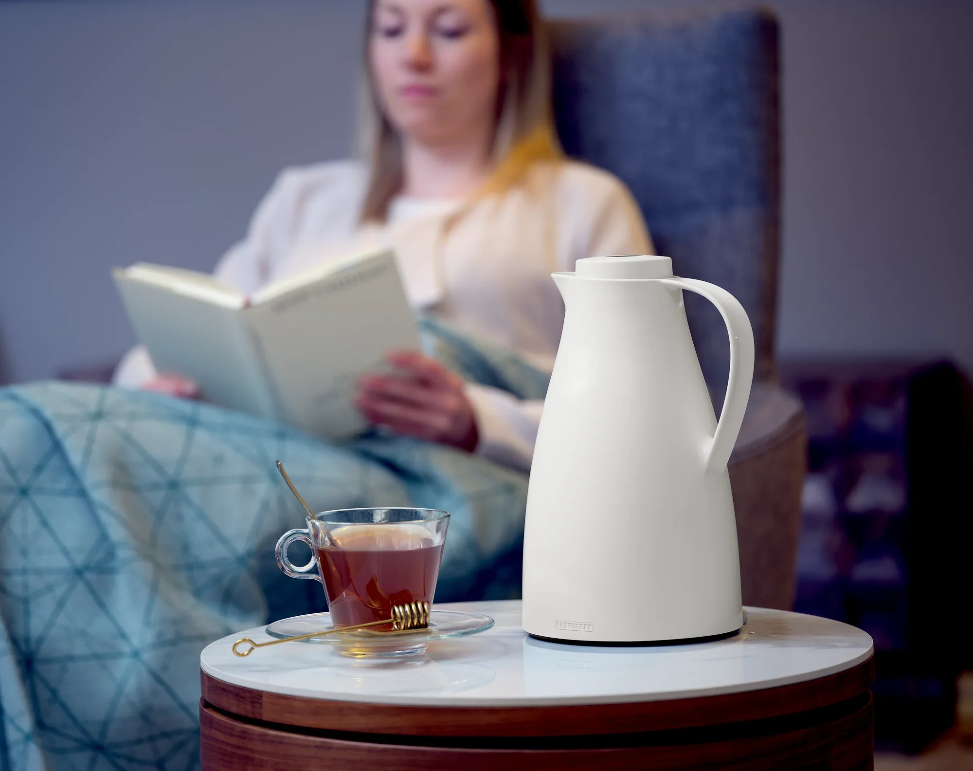 Een witte vacuümkan met de naam Harmonic staat op een ronde tafel naast een kopje thee. Op de achtergrond zit een vrouw ontspannen in een leunstoel en leest een boek.