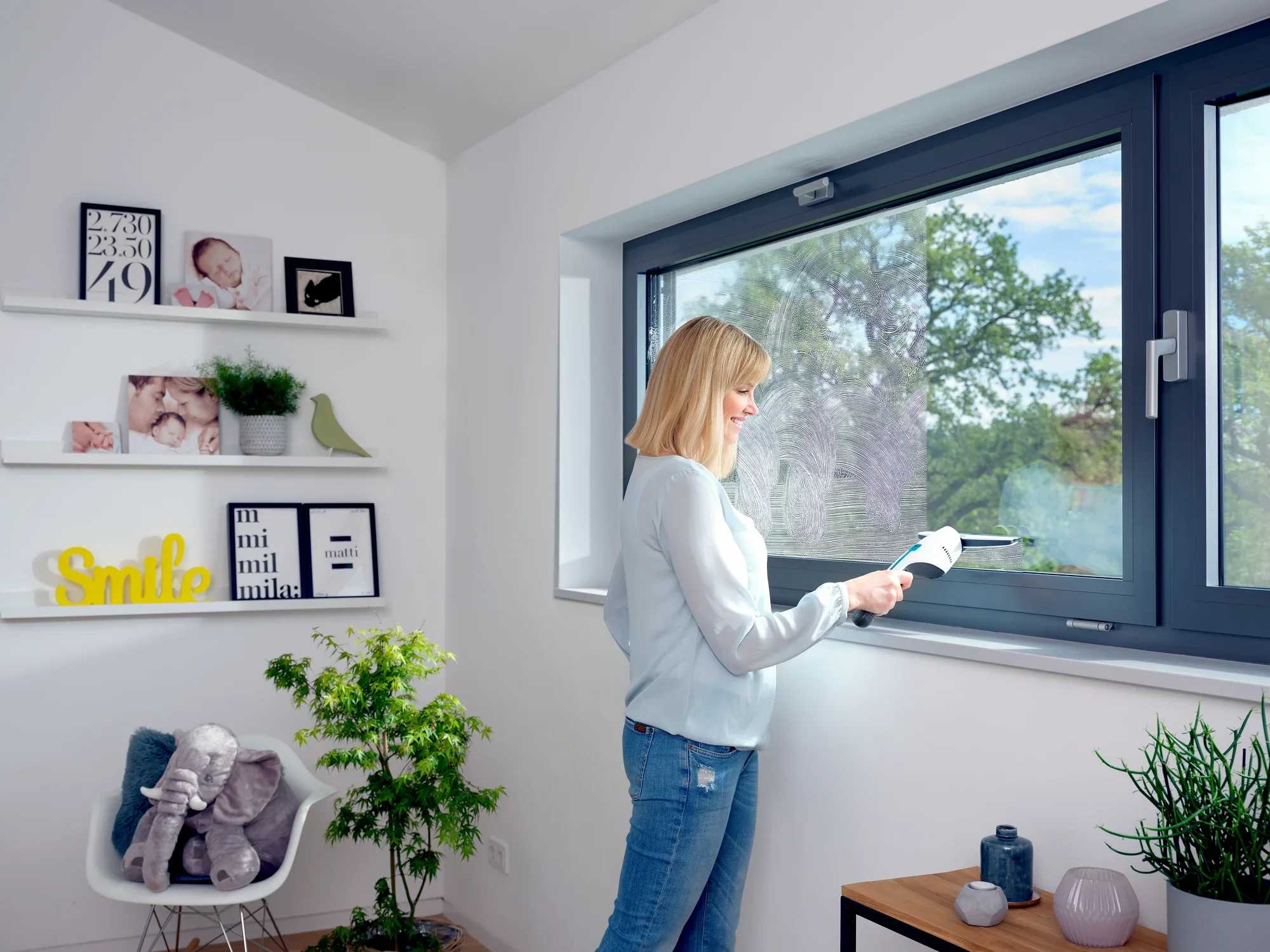 Une femme utilise le nettoyeur de vitres Nemo pour nettoyer une grande fenêtre. La pièce est lumineuse et meublée de façon moderne, avec des plantes et des éléments décoratifs.