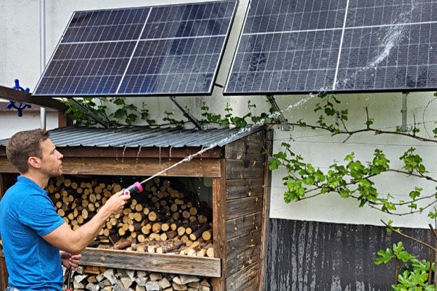 Ein Mann in blauem T-Shirt reinigt Solarpanele ueber einem Holzlager an einer weiss gestrichenen Wand mit einem Schlauch.