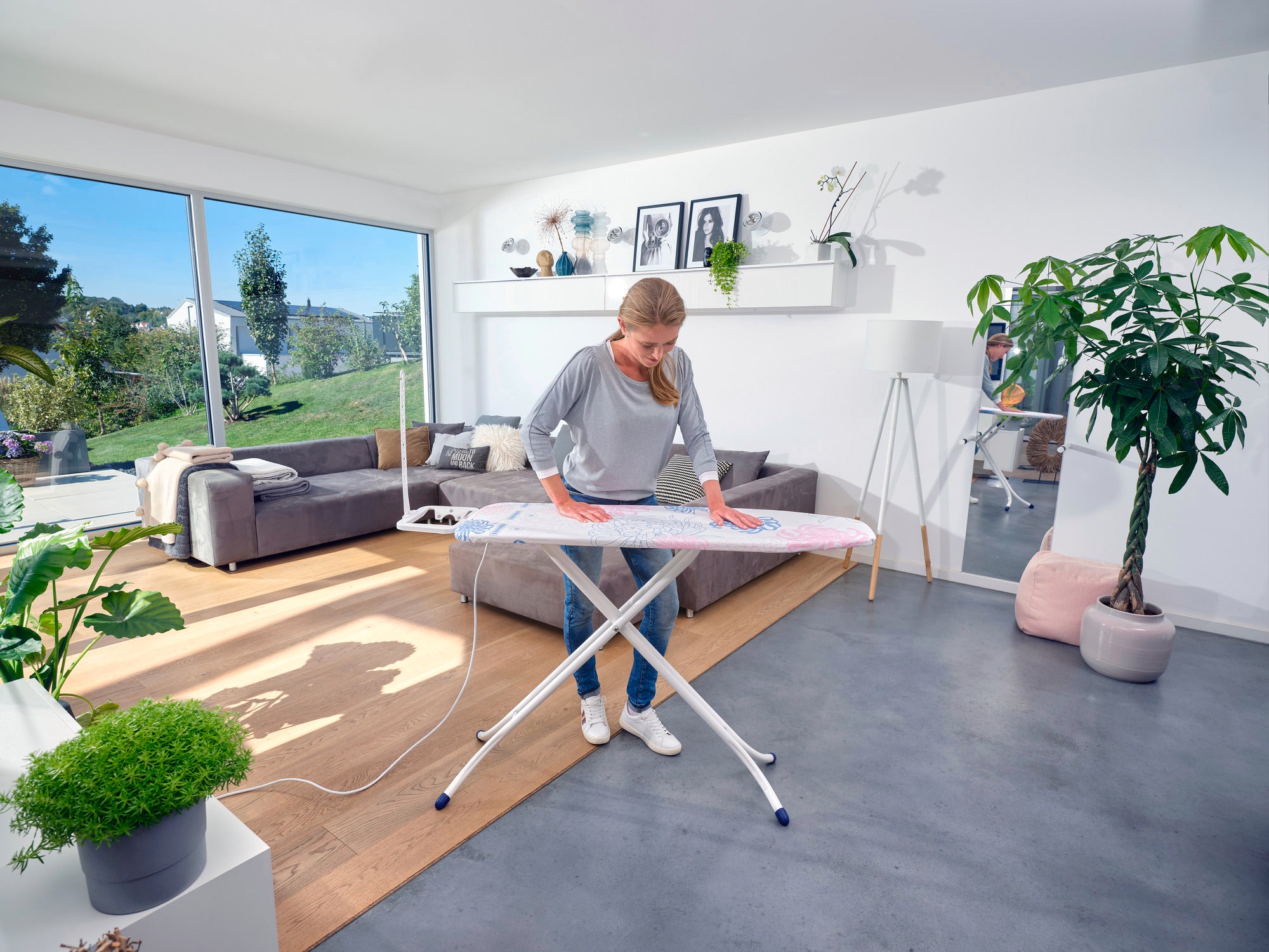 Eine Person bügelt Kleidung auf einem Bügeltisch Classic M Compact Plus in einem modernen Wohnzimmer mit großen Fenstern und Pflanzen. Der Raum ist hell und einladend gestaltet.