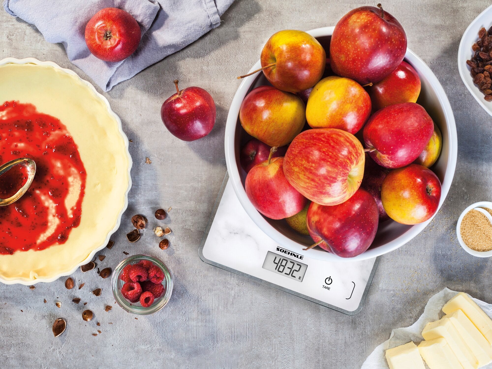 Eine digitale Küchenwaage der Marke Page Compact 300 Marble zeigt das Gewicht von Äpfeln in einer Schüssel an. Umgeben von Backzutaten wie Himbeeren und Butter.