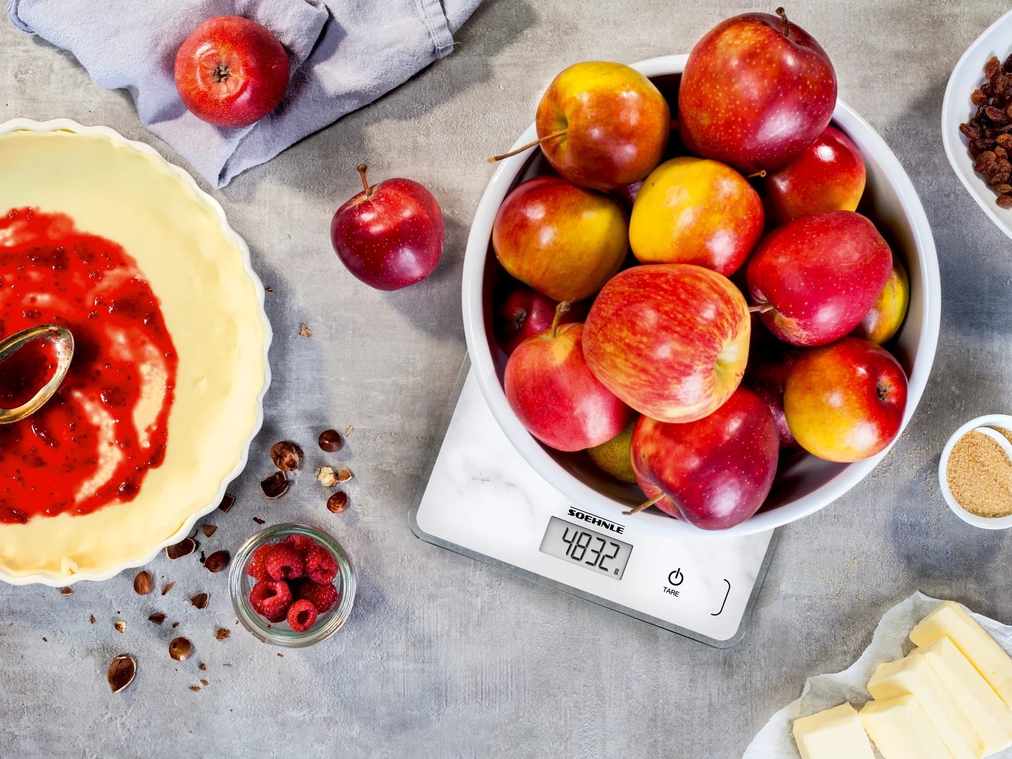 A Page Compact 300 Marble digital kitchen scale displays the weight of apples in a bowl. Surrounded by baking ingredients such as raspberries and butter.