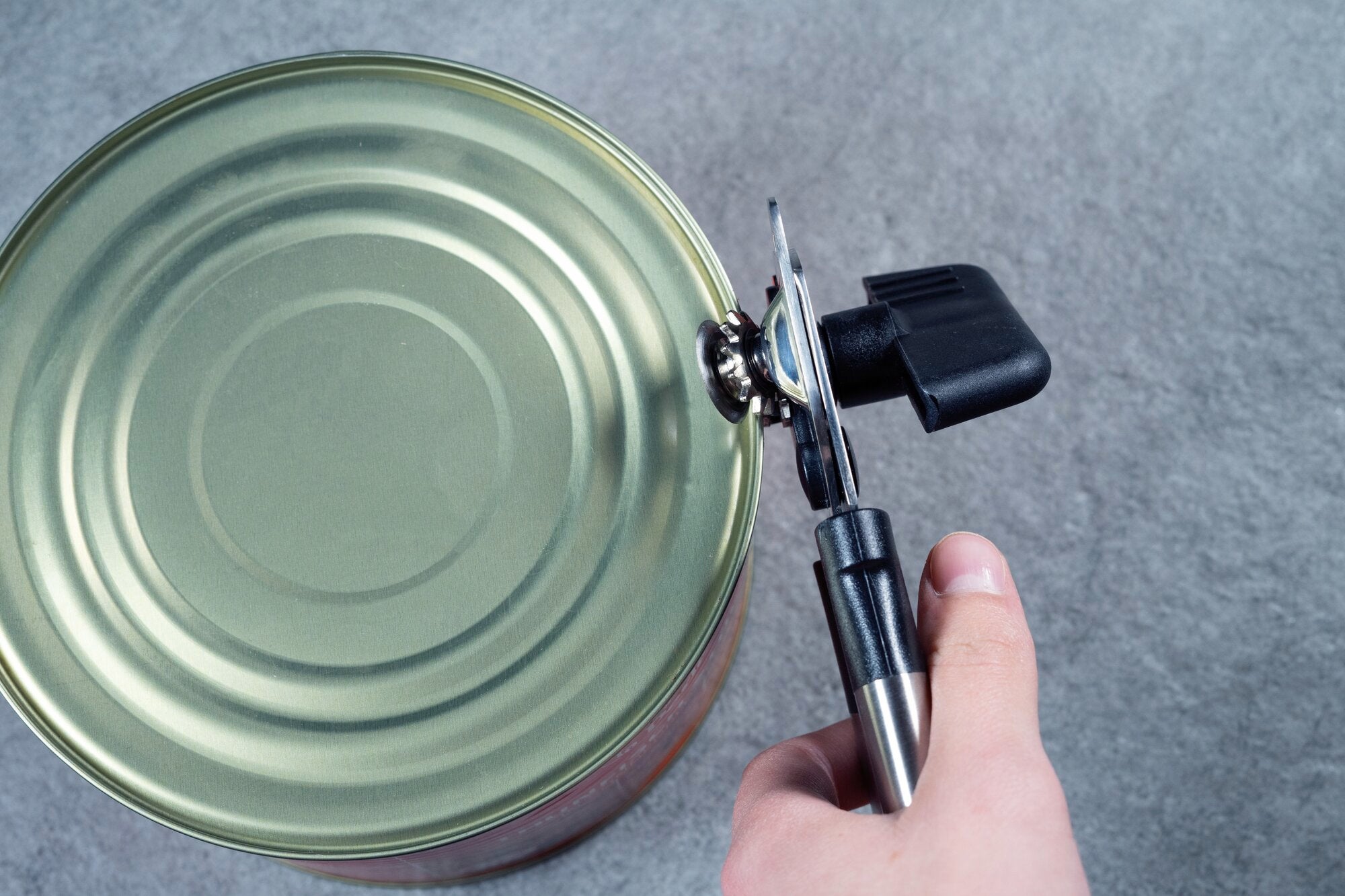 A can opener is used to open the lid of a large tin can. The hand holds the ergonomic handle of the can opener while the blade cuts the edge of the can.