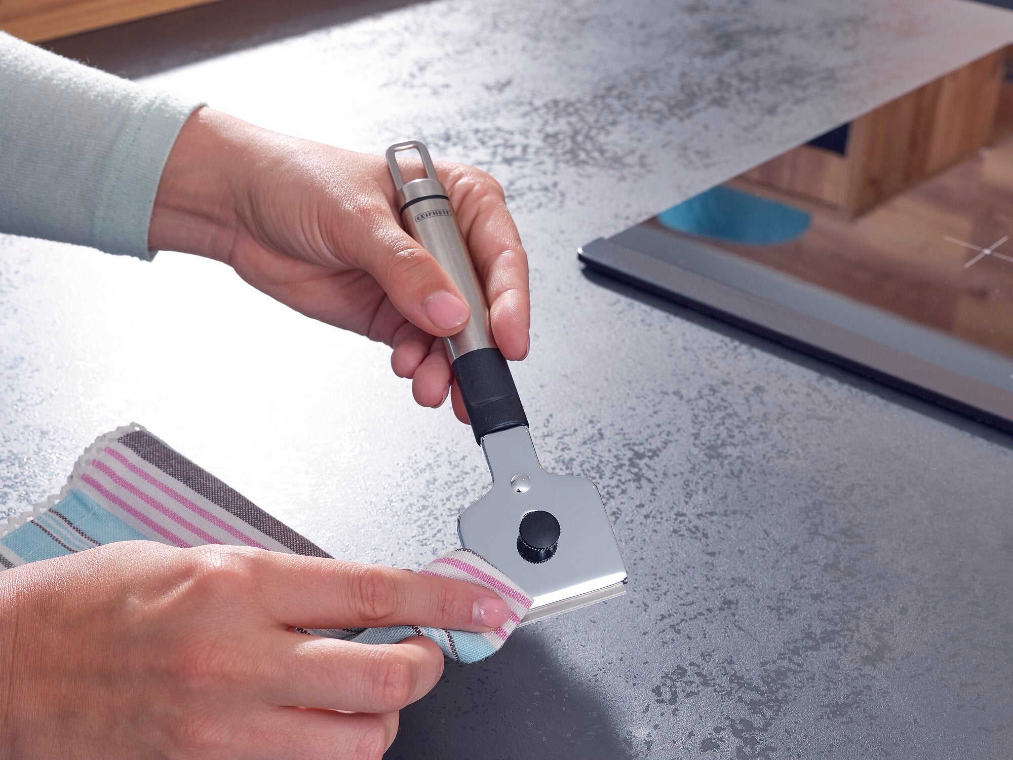 Eine Person verwendet einen Kochfeld-Schaber, um ein Kochfeld zu reinigen. Der Schaber hat einen ergonomischen Griff und wird mit einem Tuch benutzt, um Schmutz zu entfernen.