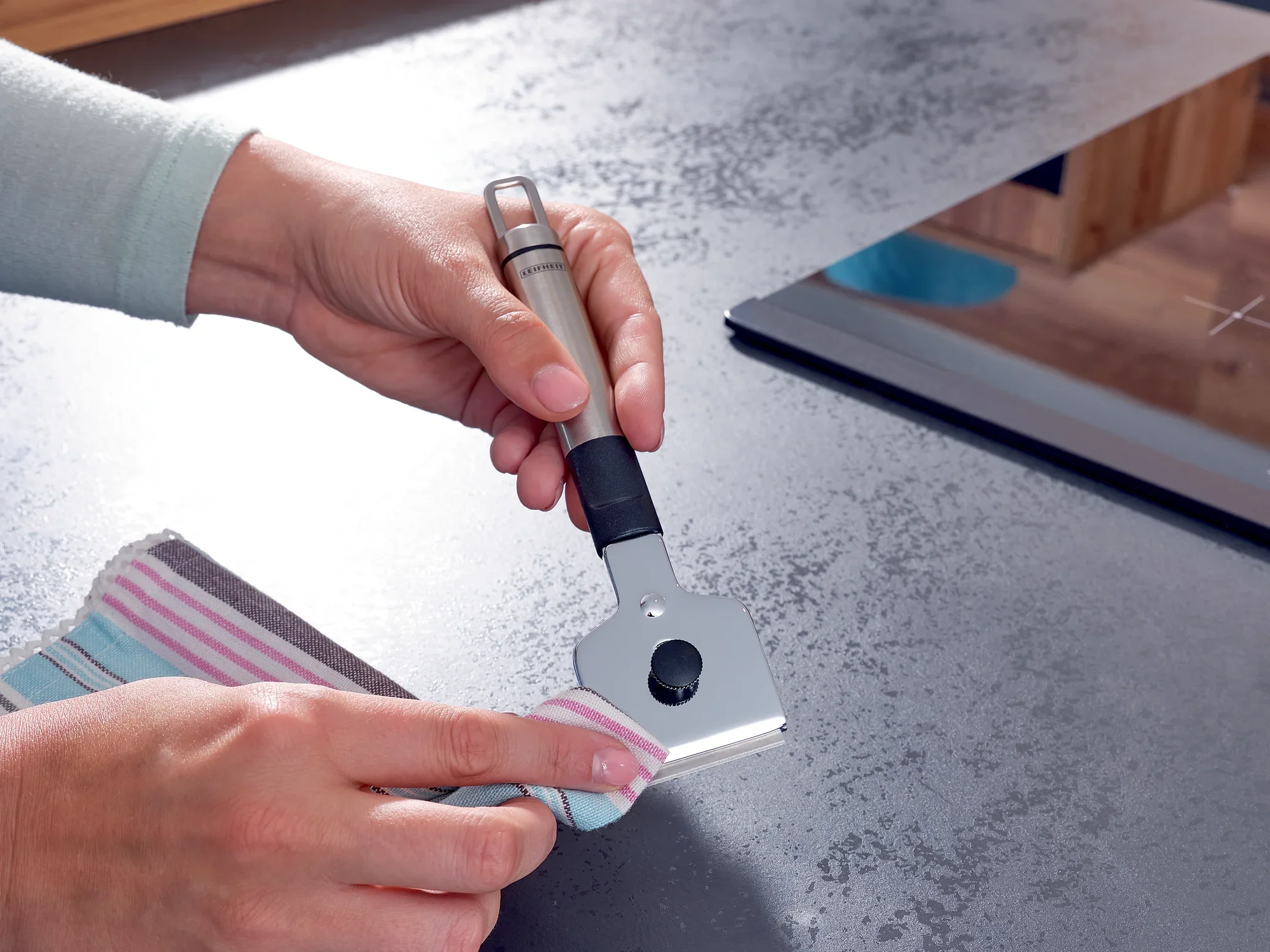 Jedna osoba używa skrobaka do czyszczenia płyty kuchennej. Skrobak ma ergonomiczny uchwyt i jest używany ze ściereczką do usuwania zabrudzeń.