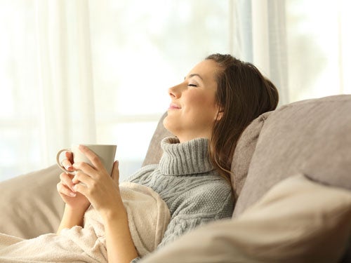 Frau sitzt entspannt auf Sofa mit grauem Pulli, haelt Tasse und geniesst Moment mit geschlossenen Augen, Tageslicht durch Fenster.