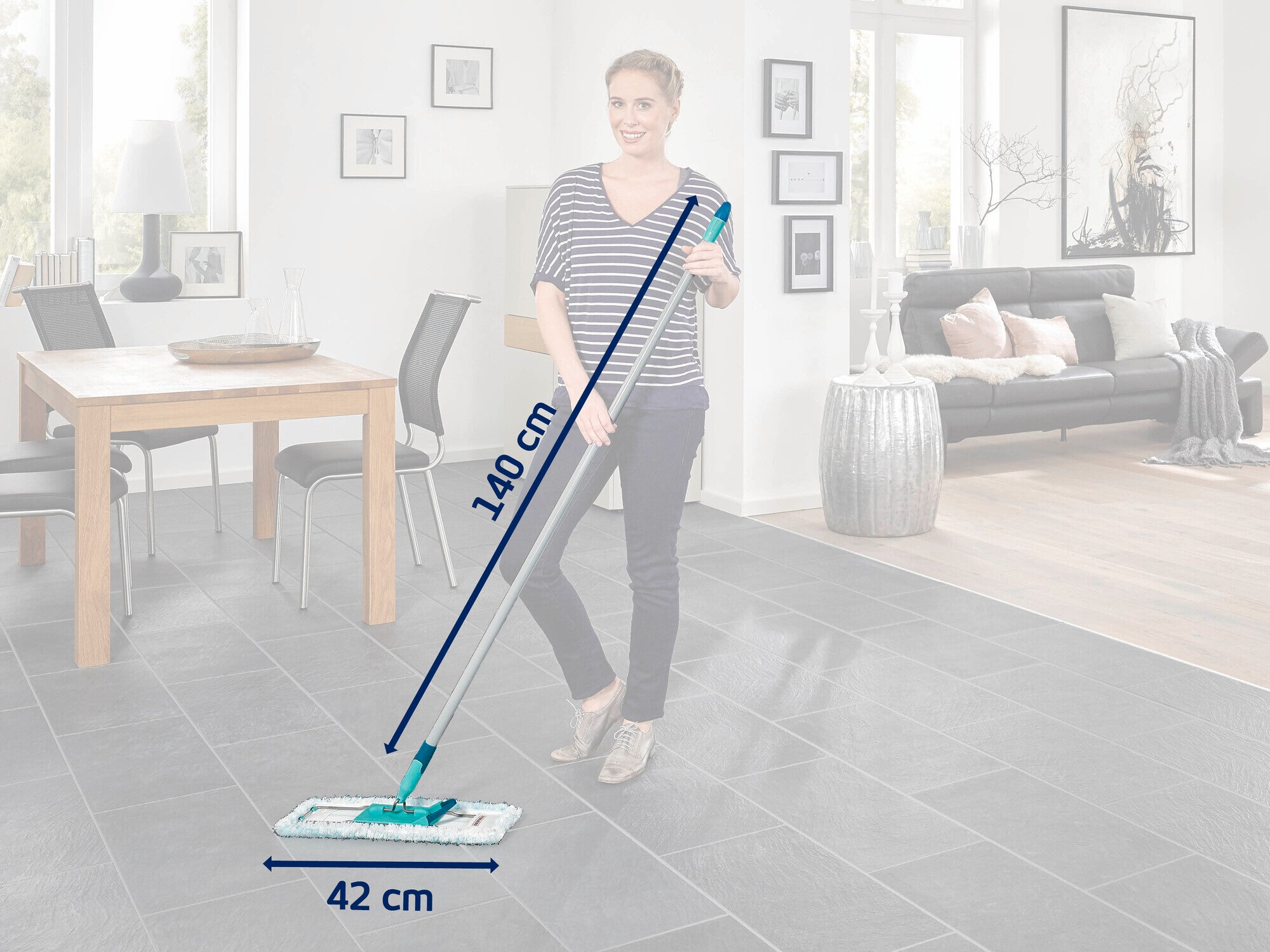 A woman cleans the floor of a modern living room with a Leifheit floor mop. The mop is 140 cm long and 42 cm wide. A dining table and sofa can be seen in the background.