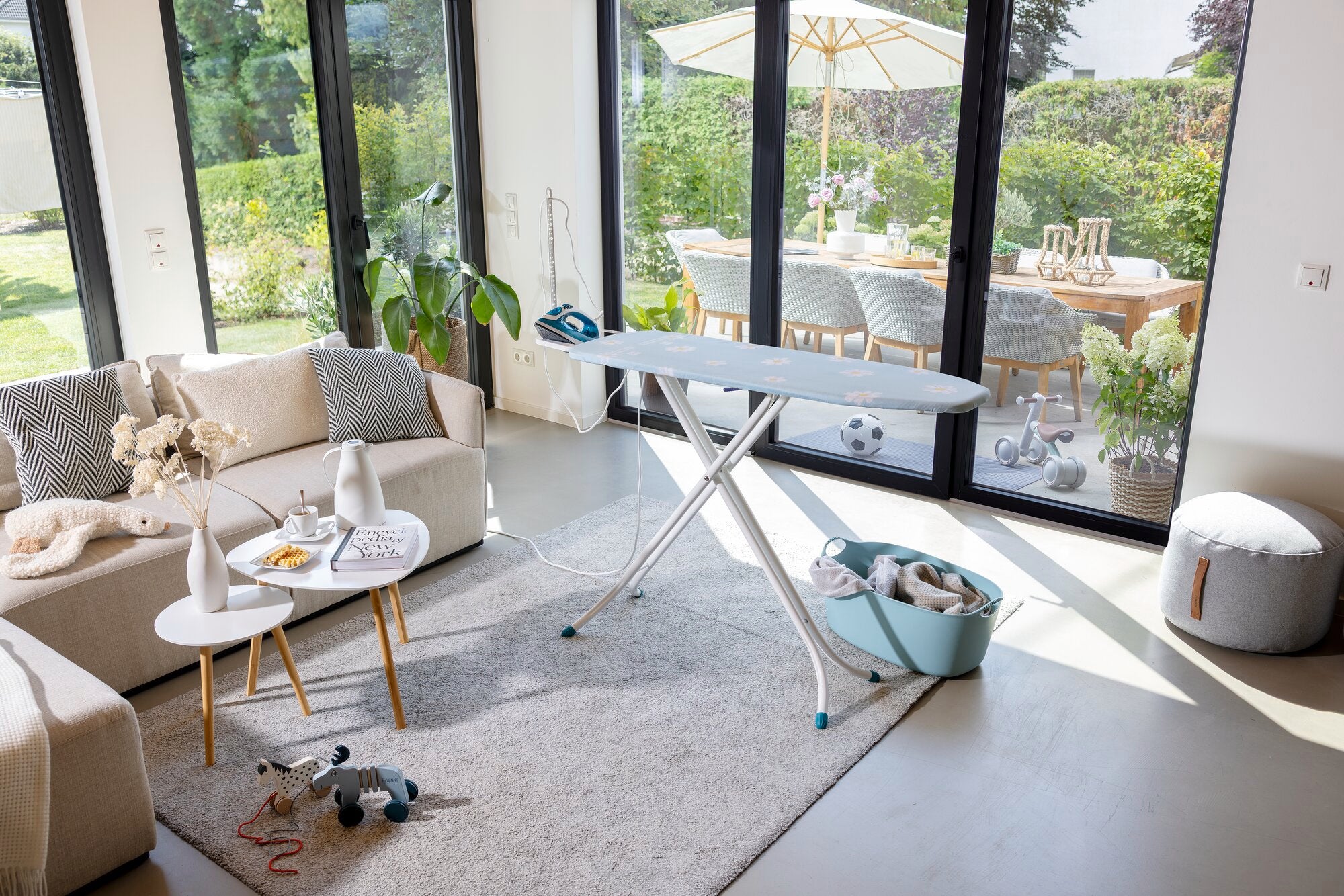 Bügelbrett Leifheit Classic M Compact Plus in einem modernen Wohnzimmer mit großen Fenstern. Das Brett steht auf einem hellen Teppich, umgeben von stilvollen Möbeln und Dekorationen.