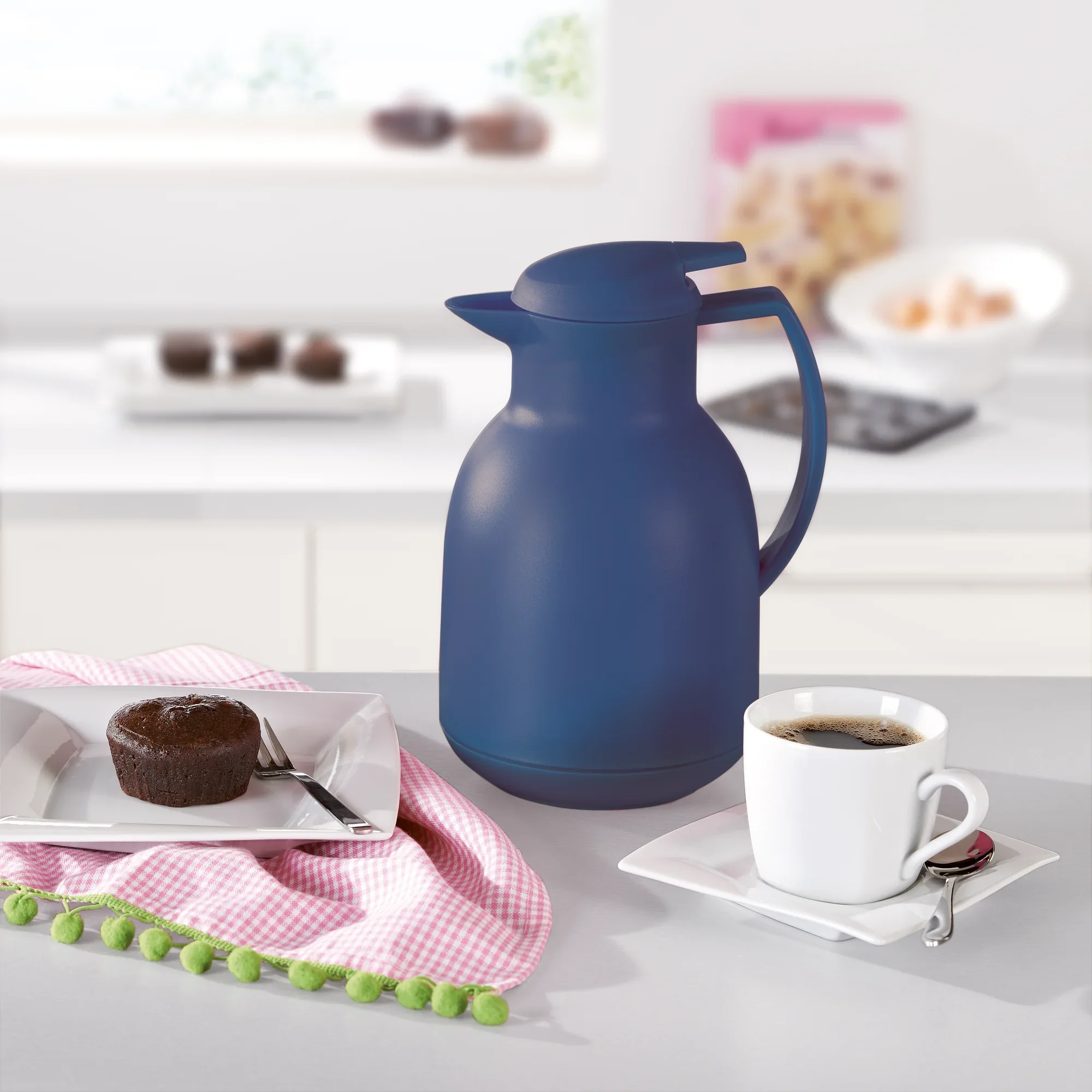 Eine dunkelblaue Isolierkanne "Bolero" mit 1000 ml Fassungsvermögen steht auf einem Tisch. Daneben eine Tasse Kaffee und ein Schokoladenmuffin auf einem Teller mit rosa Serviette.