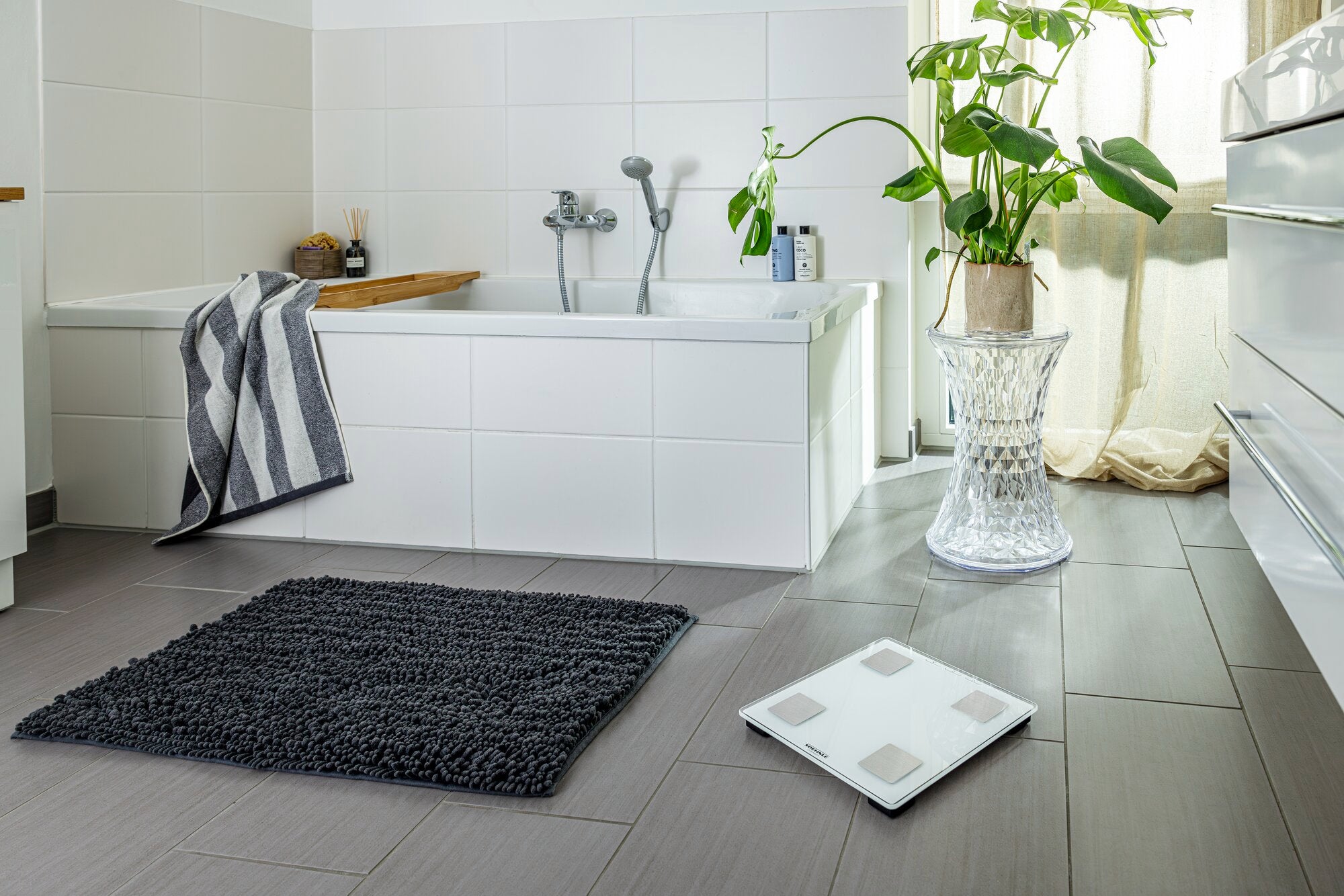 Shape Sense Connect Lite digital bathroom scales in a modern bathroom. The scales are on a grey tiled floor next to a black rug and a bathtub.