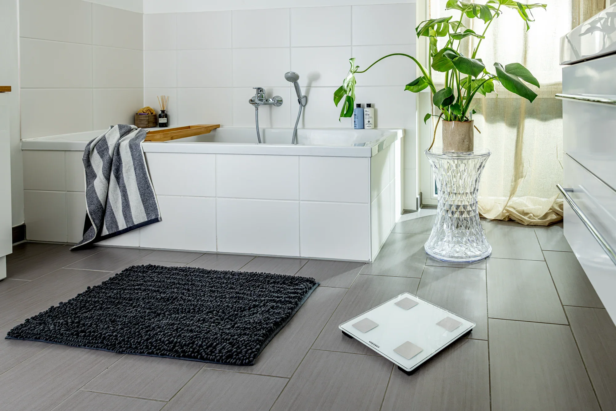 Cyfrowa waga Shape Sense Connect Lite w nowoczesnej łazience. Waga leży na szarej podłodze wyłożonej kafelkami obok czarnego dywanu i wanny.