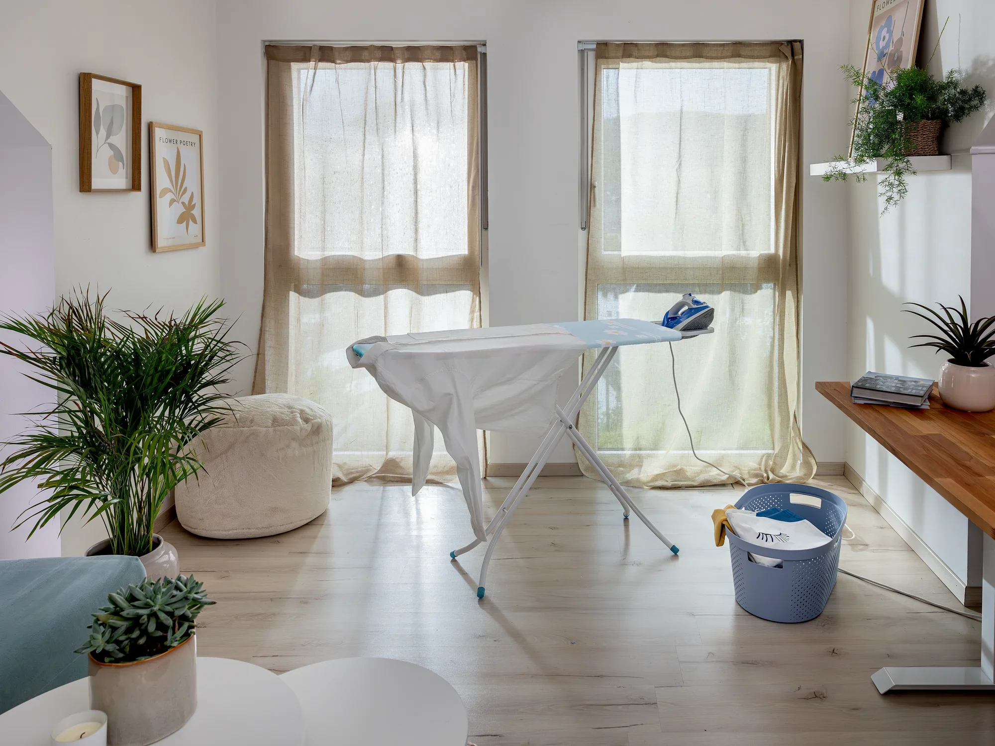 Bügelbrett Leifheit Classic M Basic in einem hellen Raum mit großen Fenstern. Das Bügelbrett steht in der Mitte, darauf ein Bügeleisen. Pflanzen und Dekorationen umgeben den Raum.