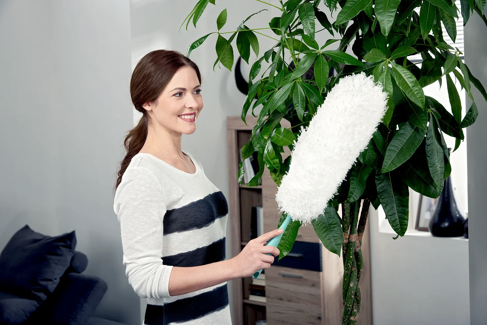 Una mujer utiliza el plumero para limpiar las hojas de una gran planta de interior. El plumero es blanco y tiene un mango verde, ideal para una limpieza suave.