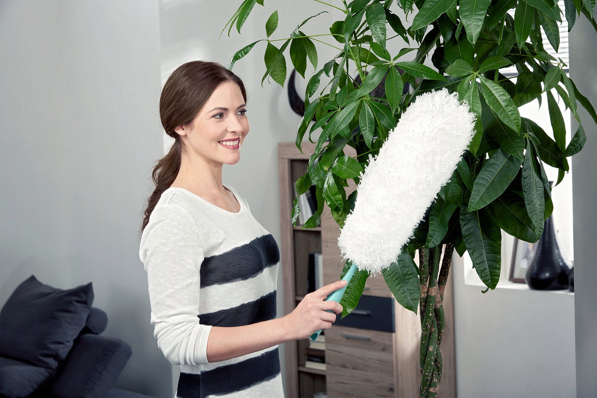 A woman uses a feather duster to clean the leaves of a large houseplant. The feather duster is white and has a green handle, ideal for gentle cleaning.